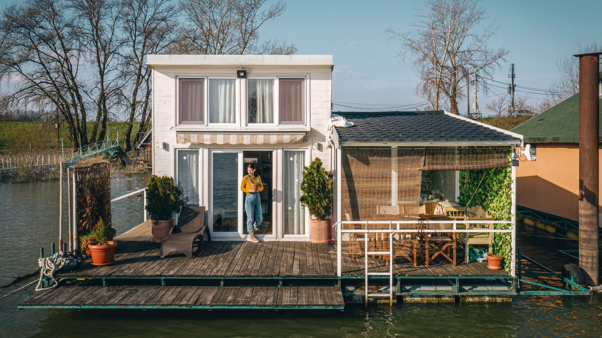 Weißes zweistöckiges Hausboot mit Holzterrasse, Pflanzenkübeln und überdachter Sitzecke auf einem Fluss bei Sonnenschein.