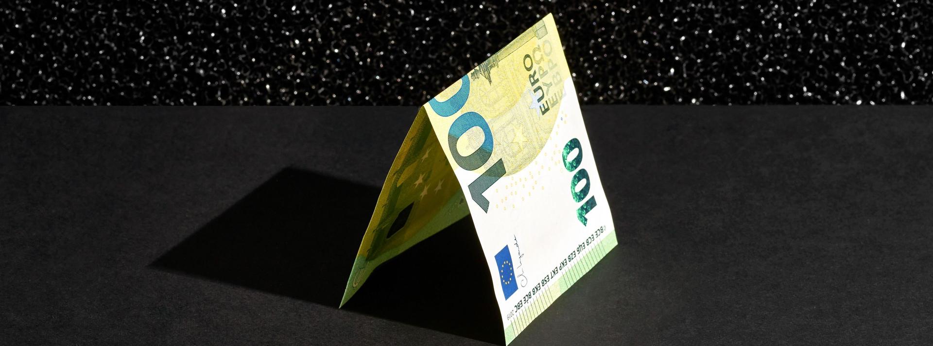 Ein 100-Euro Schein liegt gefaltet auf einem schwarzem Hintergrund.
