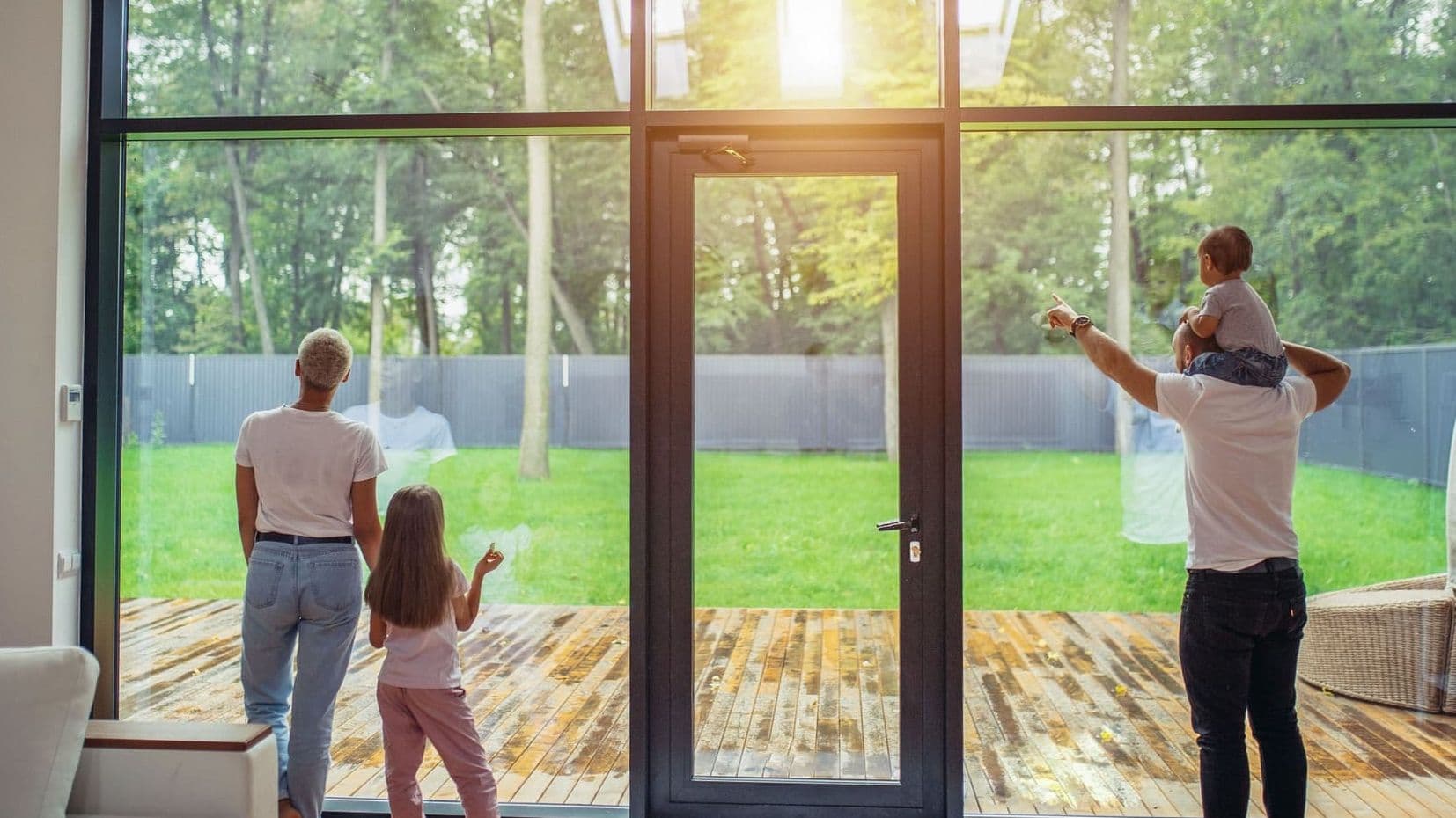 Familie steht in einem modernen Wohnzimmer vor großer Glasfront und blickt auf den grünen Garten, Vater trägt Kleinkind auf den Schultern