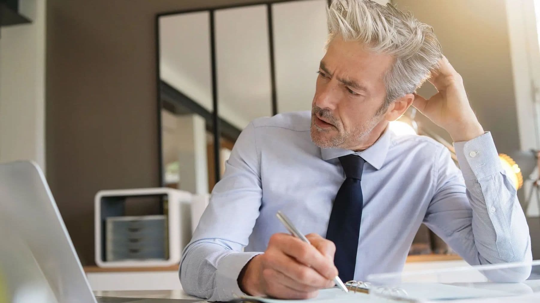 Mann mit grauen Haaren sitzt am Schreibtisch, schaut nachdenklich auf Unterlagen und arbeitet am Laptop in einem Büro