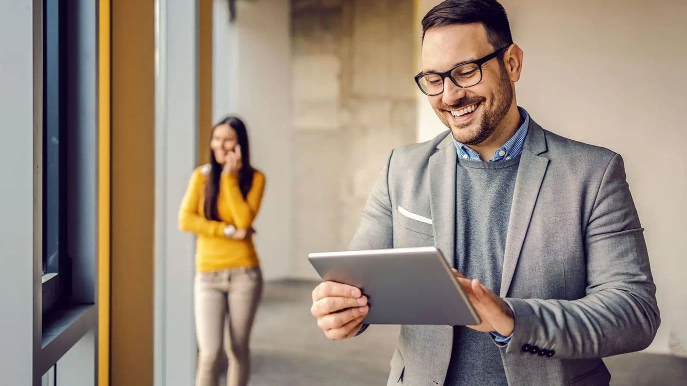 Mann in grauem Anzug betrachtet lächelnd ein Tablet, im Hintergrund steht eine Frau in gelbem Pullover telefonierend in einem modernen Gebäude