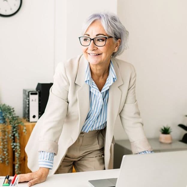 Ältere Frau mit grauen Haaren und Brille steht lächelnd an einem Schreibtisch mit Laptop in modernem Büroumfeld