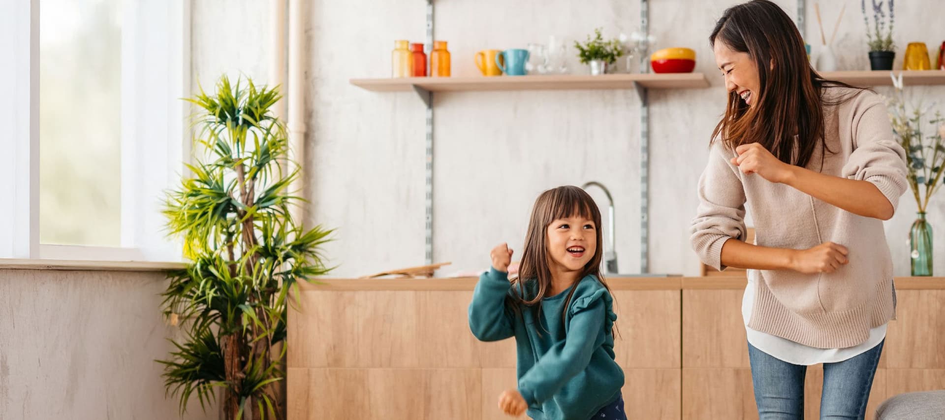 Frau tanzt lachend mit jungem Kind im hellen Wohnzimmer mit Küche im Hintergrund