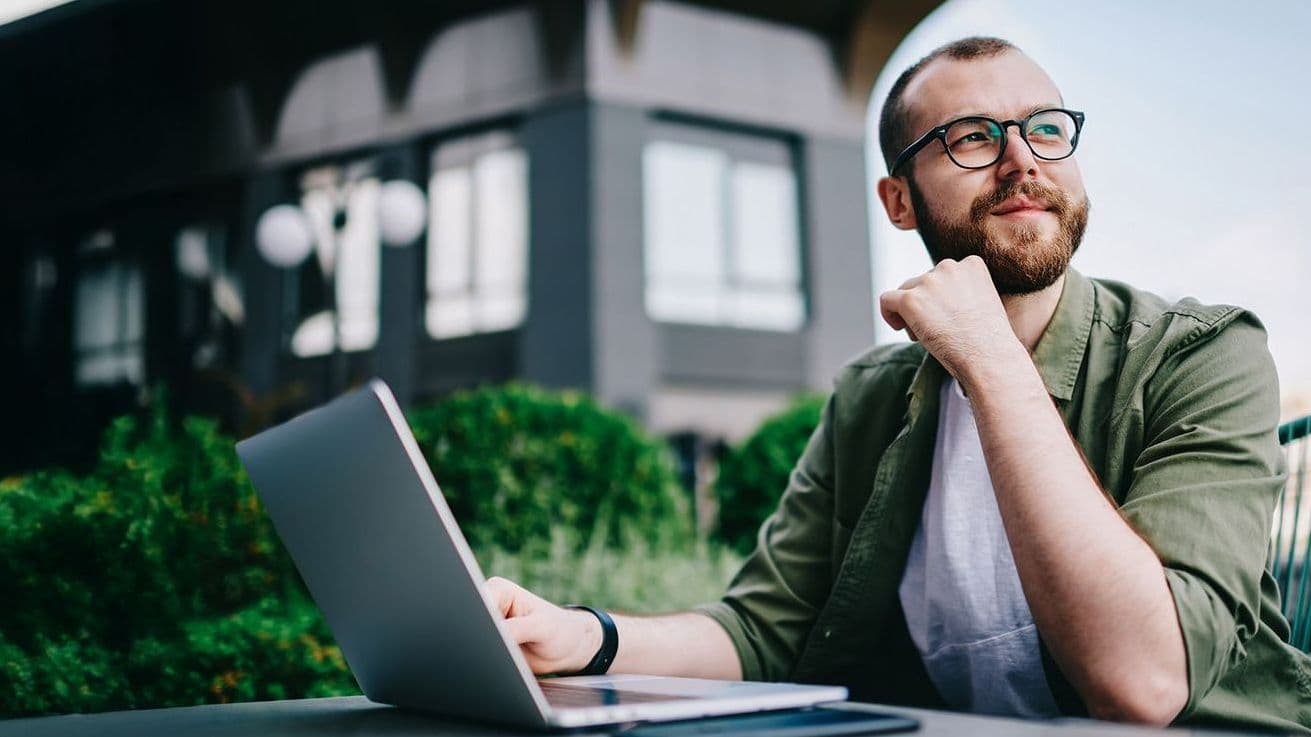 Person mit Laptop sitzt im Freien an einem Tisch, blickt nachdenklich in die Ferne, modernes Gebäude und grüne Pflanzen im Hintergrund