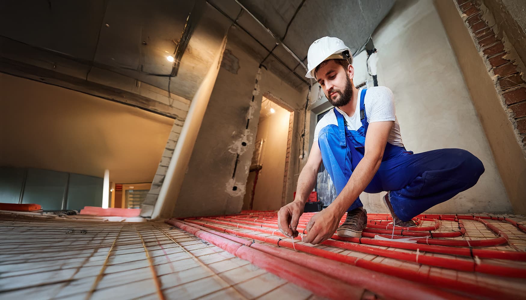 Handwerker verlegt Fußbodenheizung mit roten Heizrohren in unfertigem Innenraum während Renovierung