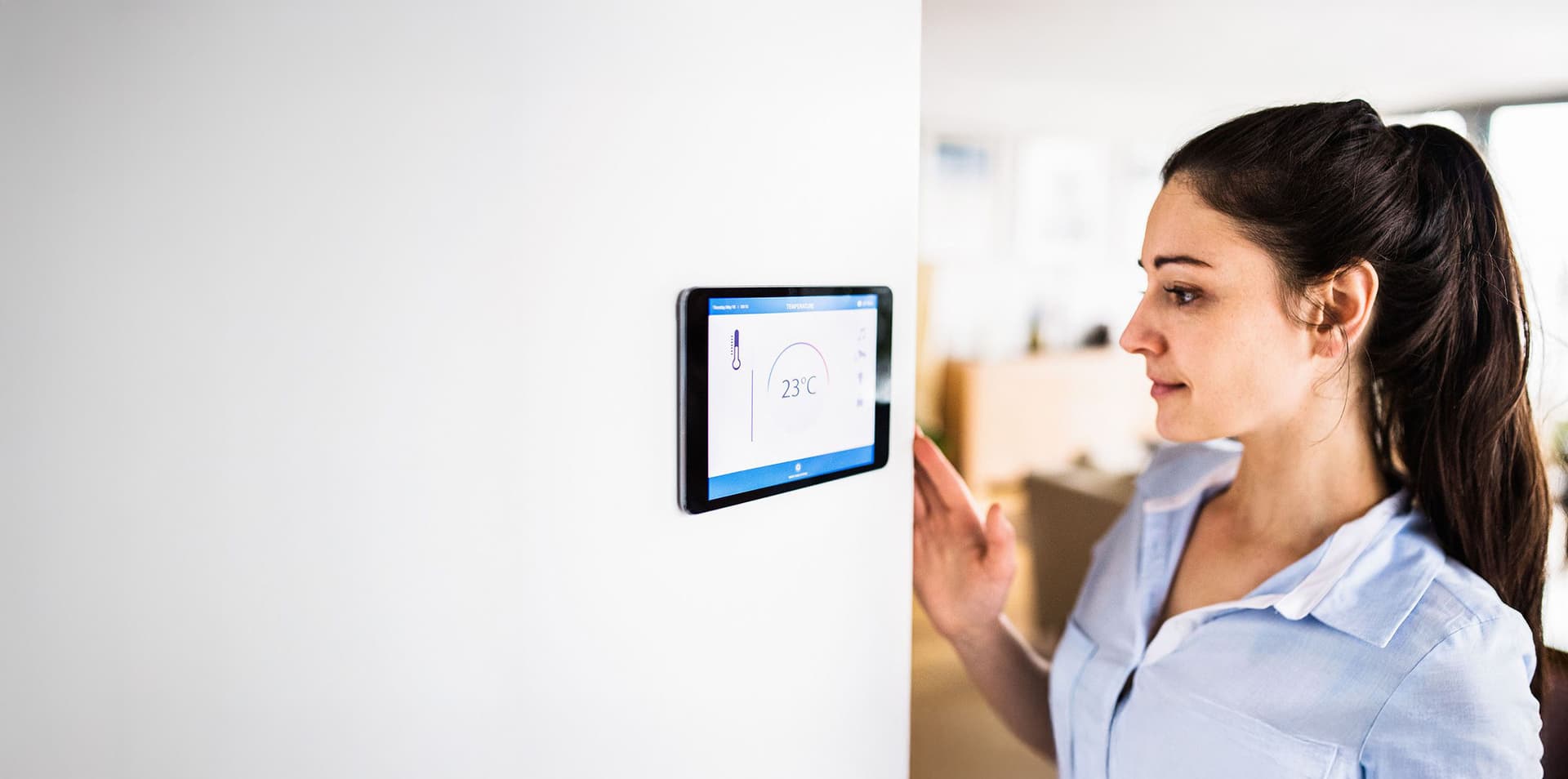 Frau bedient ein Smart-Home-Thermostat an der Wand; Display zeigt 23°C.
