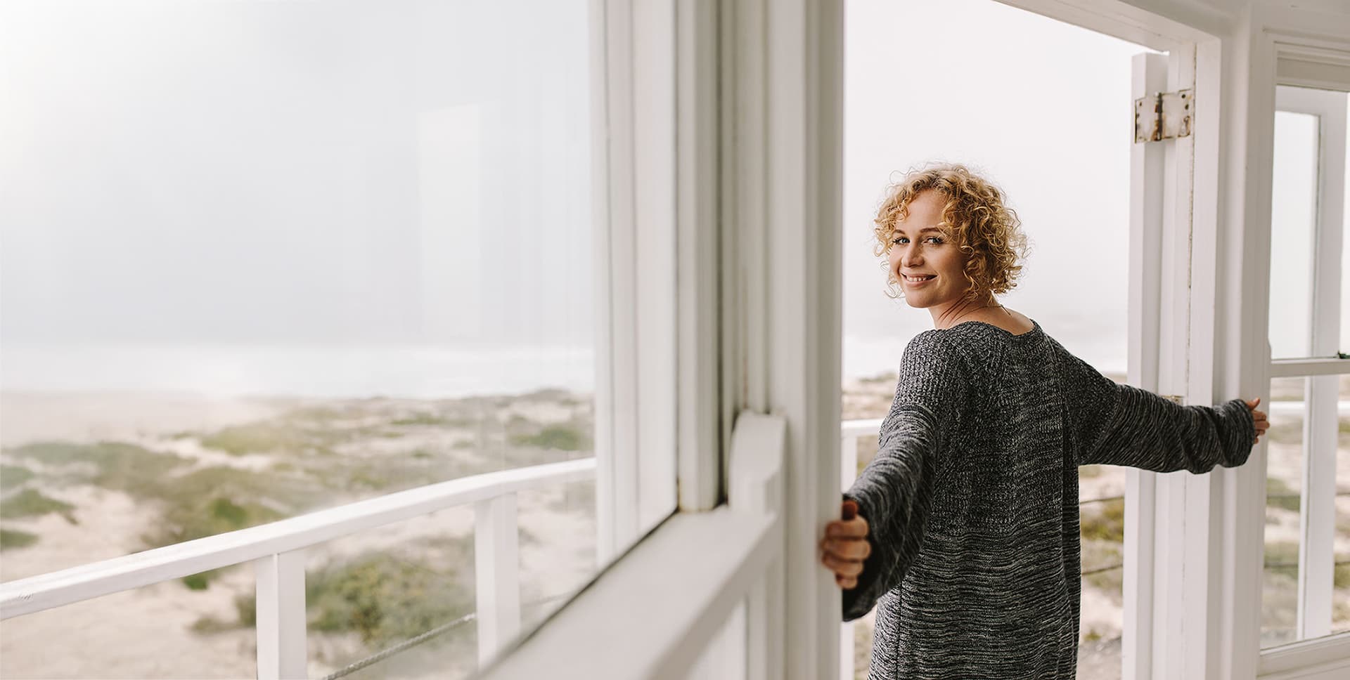 Frau steht in einem Strandhaus, hält die Tür offen und blickt zum Meer.