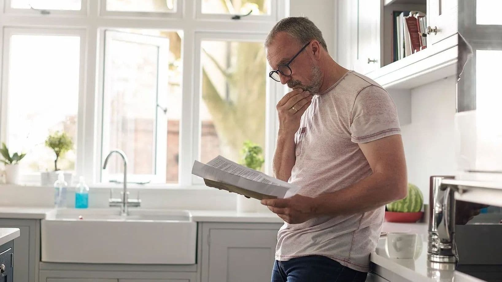 Mann mittleren Alters liest Dokument in heller Küche und wirkt nachdenklich