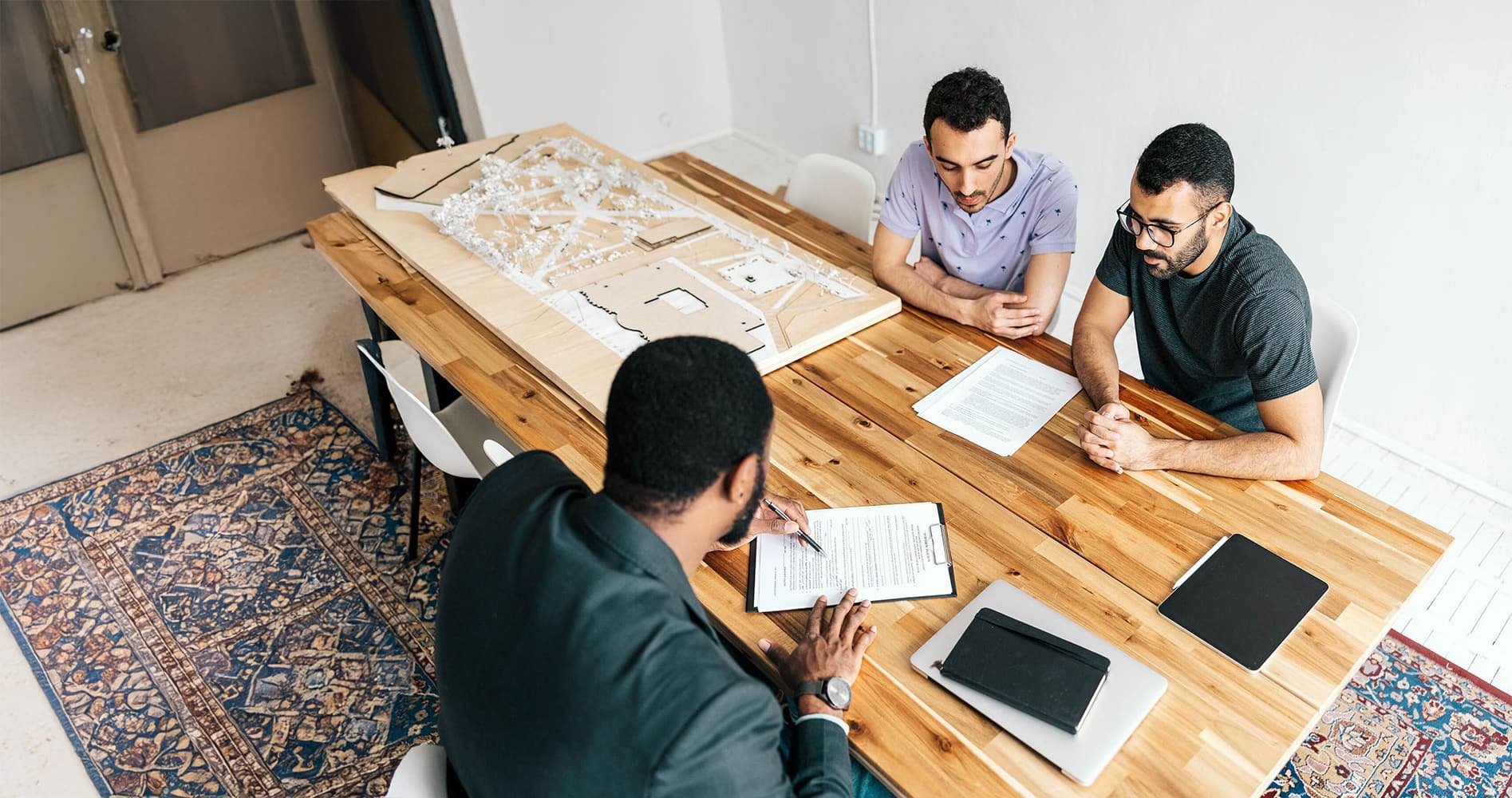 Zwei Männer sitzen gegenüber einem Berater an einem Holztisch und unterschreiben Vertragsdokumente bei einer Beratungssituation.