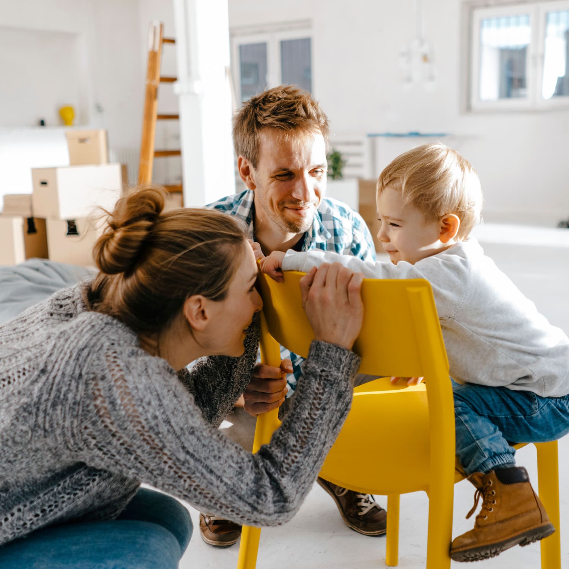 Junge Familie in einem modernen noch nicht fertig eingerichteten Raum. Die Mutter hockt vor einem gelben Stuhl und versteckt ihr Gesicht vor dem Kleinkind, was auf dem Stuhl sitzt.