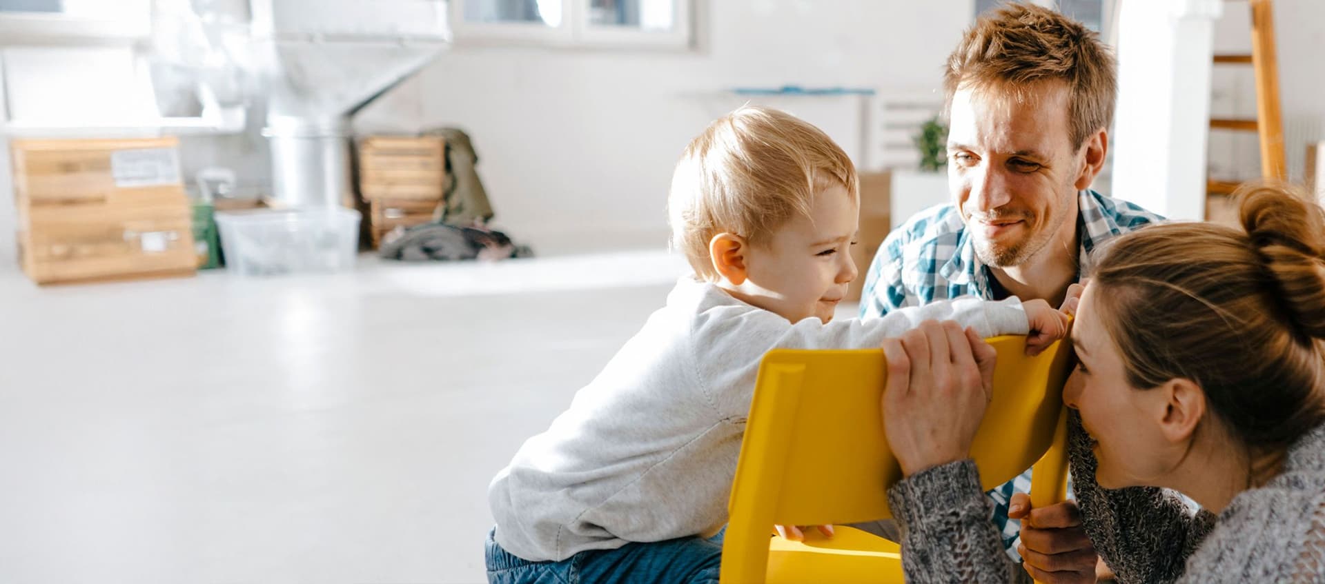 Junge Familie in einem modernen noch nicht fertig eingerichteten Raum. Die Mutter hockt vor einem gelben Stuhl und versteckt ihr Gesicht vor dem Kleinkind, was auf dem Stuhl sitzt.