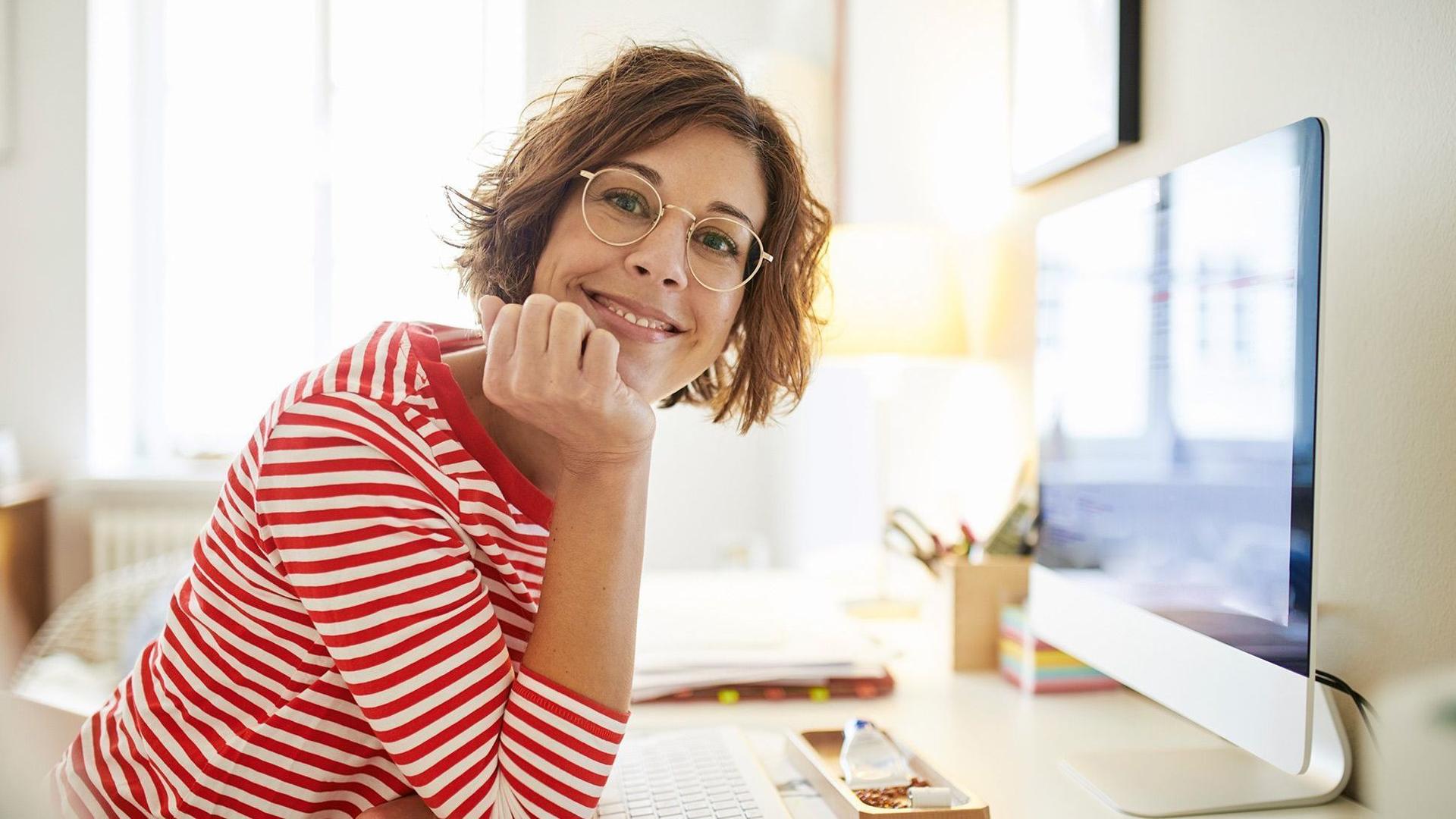 Eine junge Frau sitzt lächelnd an ihrem Schreibtisch vor einem Arbeitsbildschirm.