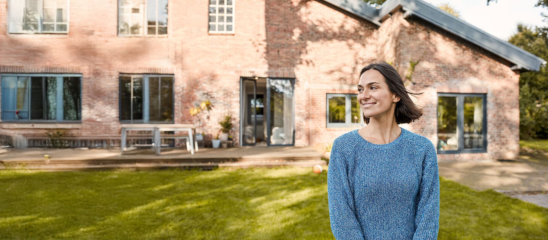 Frau steht im Garten vor einem Backsteinhaus mit Terrasse und Fenstern an einem sonnigen Tag