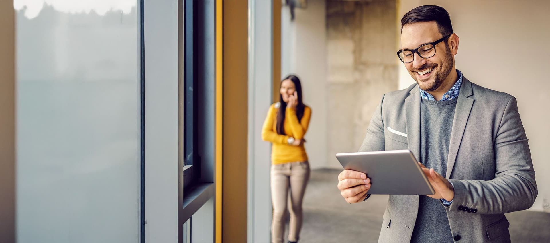 Mann mit Brille und Tablet im Büro, im Hintergrund Frau mit gelbem Pullover telefonierend