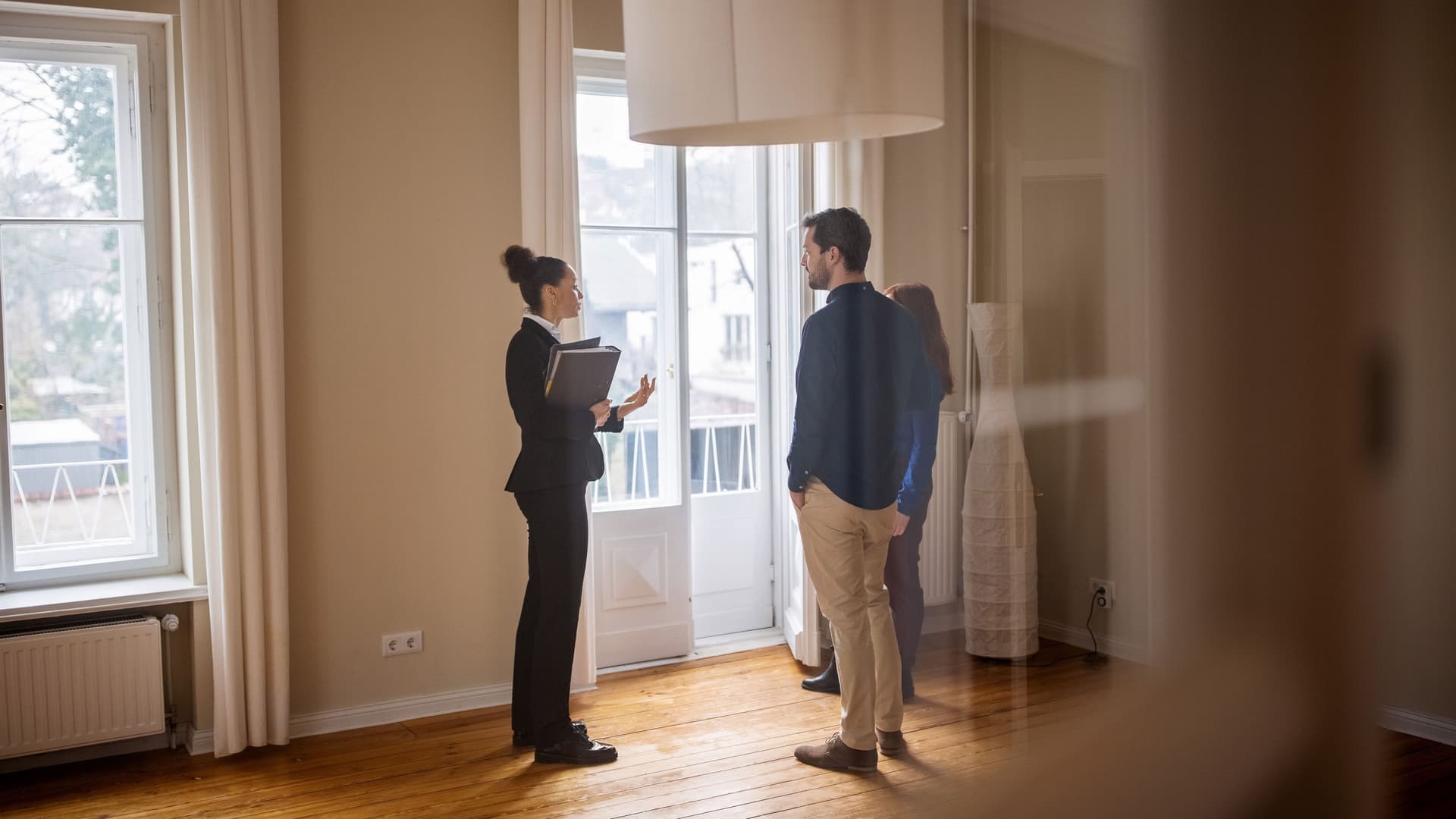 Immobilienmaklerin steht mit einem jungen Paar in einem leeren Zimmer. Sie trägt einen Hosenanzug und hat eine Mappe in der Hand.