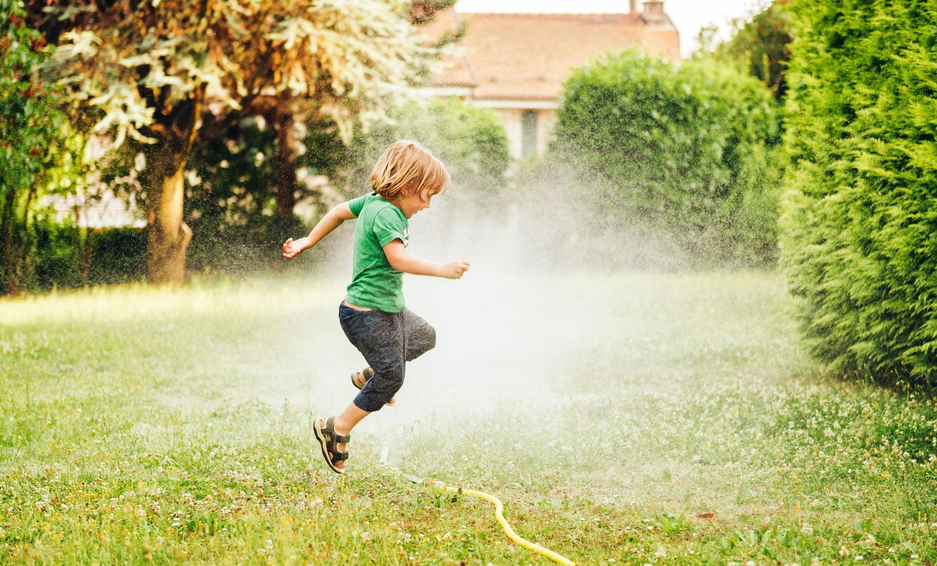Ein Kind springt im Garten über einen Rasensprenger.