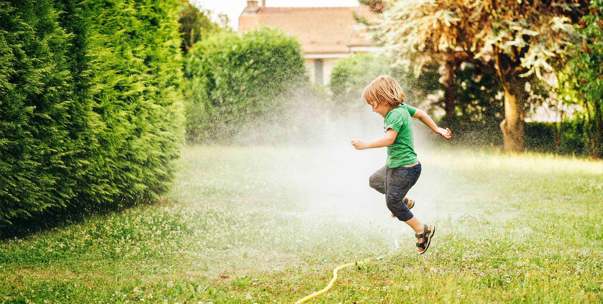 Kind spielt draußen im Garten und springt durch den Wasserstrahl eines Rasensprengers