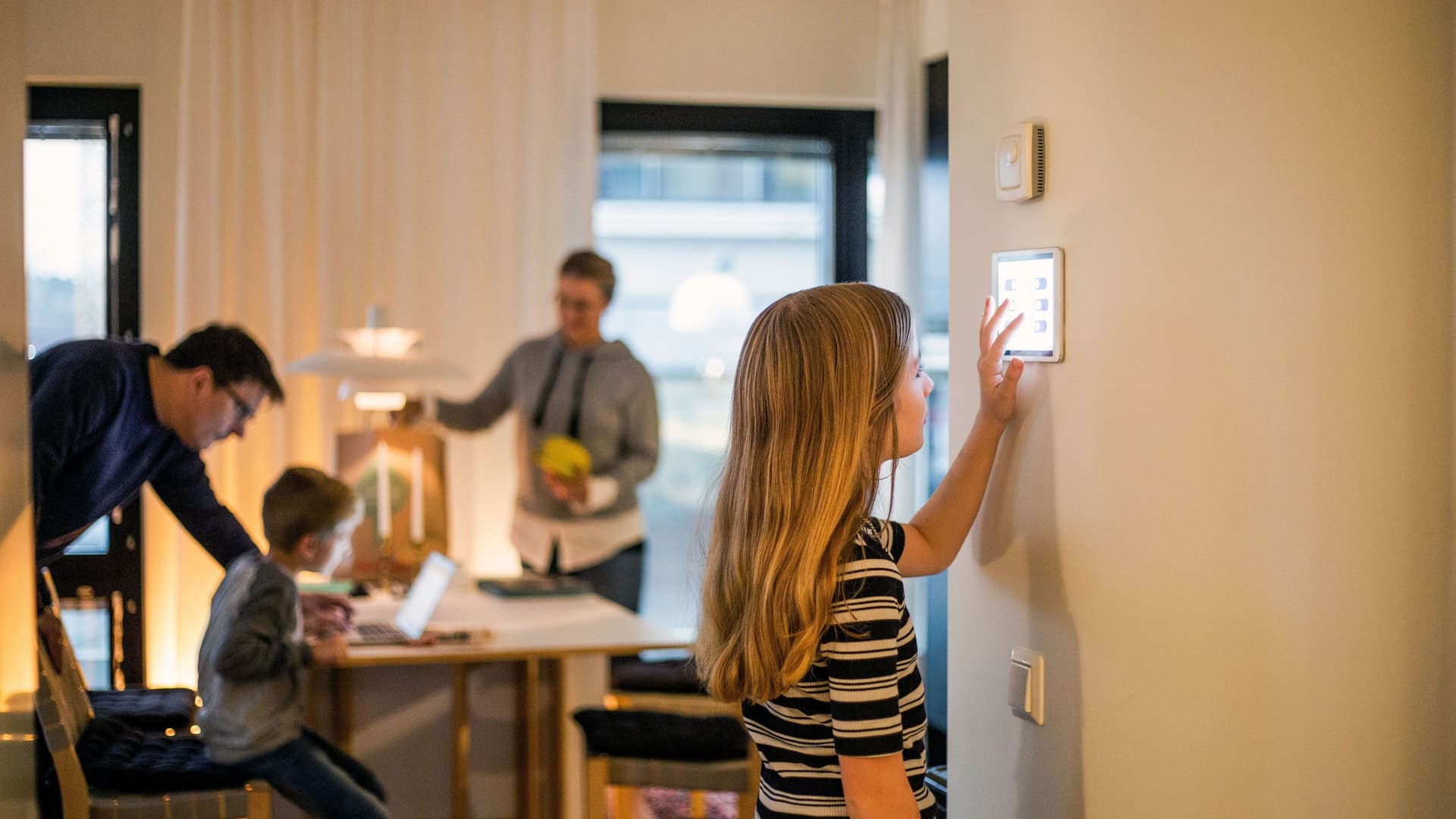 Mädchen bedient digitales Smart Home Display an der Wand. Im Hintergrund ist unscharf die Familie zu erkennen.