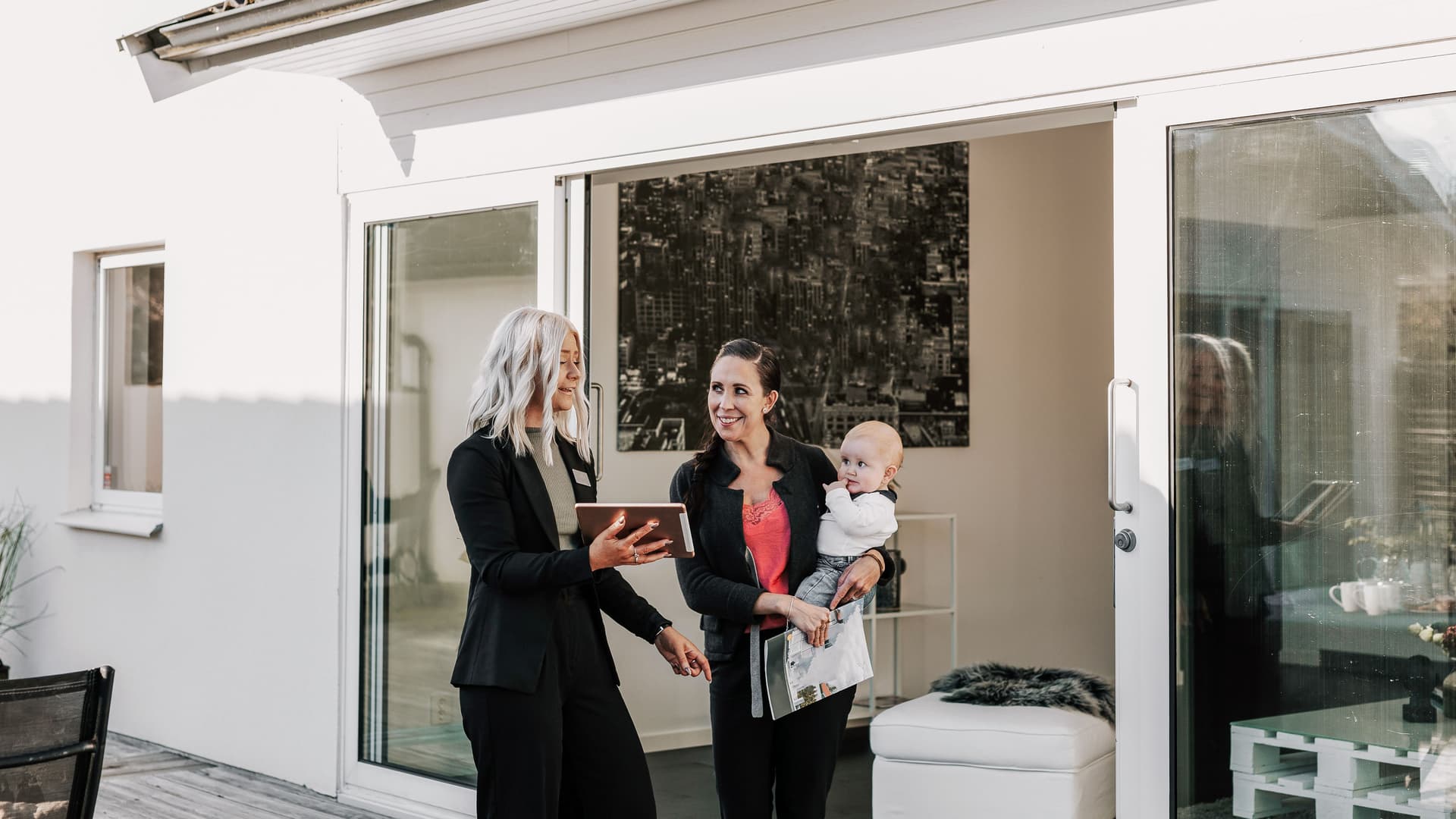 Maklerin zeigt junger Mutter mit Baby auf dem Arm eine Immobilie.