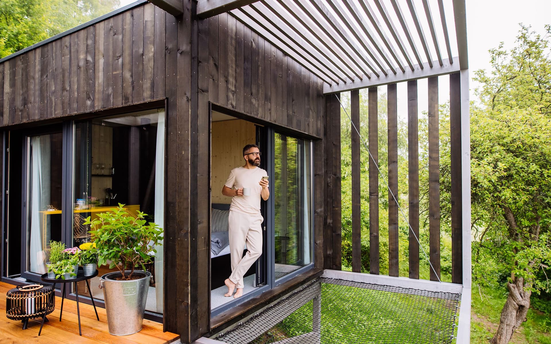 Mann mit Kaffee und Smartphone steht barfuß am Tiny-House-Fenster mit Blick ins Grüne.