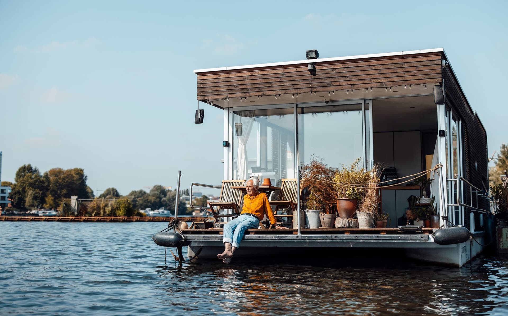 Älterer Mann sitzt entspannt auf dem Deck eines modernen Hausboots am Wasser.