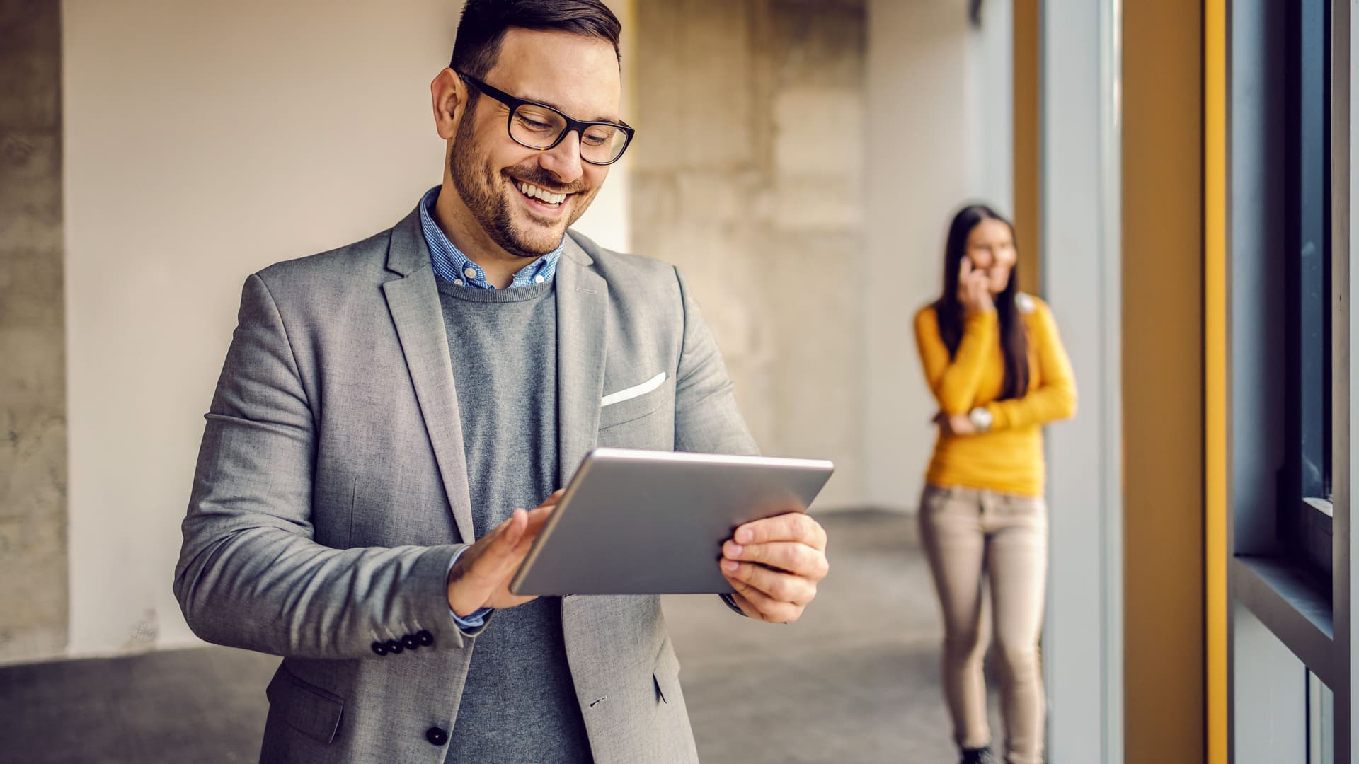 Junger Mann schaut lächelnd auf sein Tablet. Im Hintergrund steht eine telefonierende Frau.