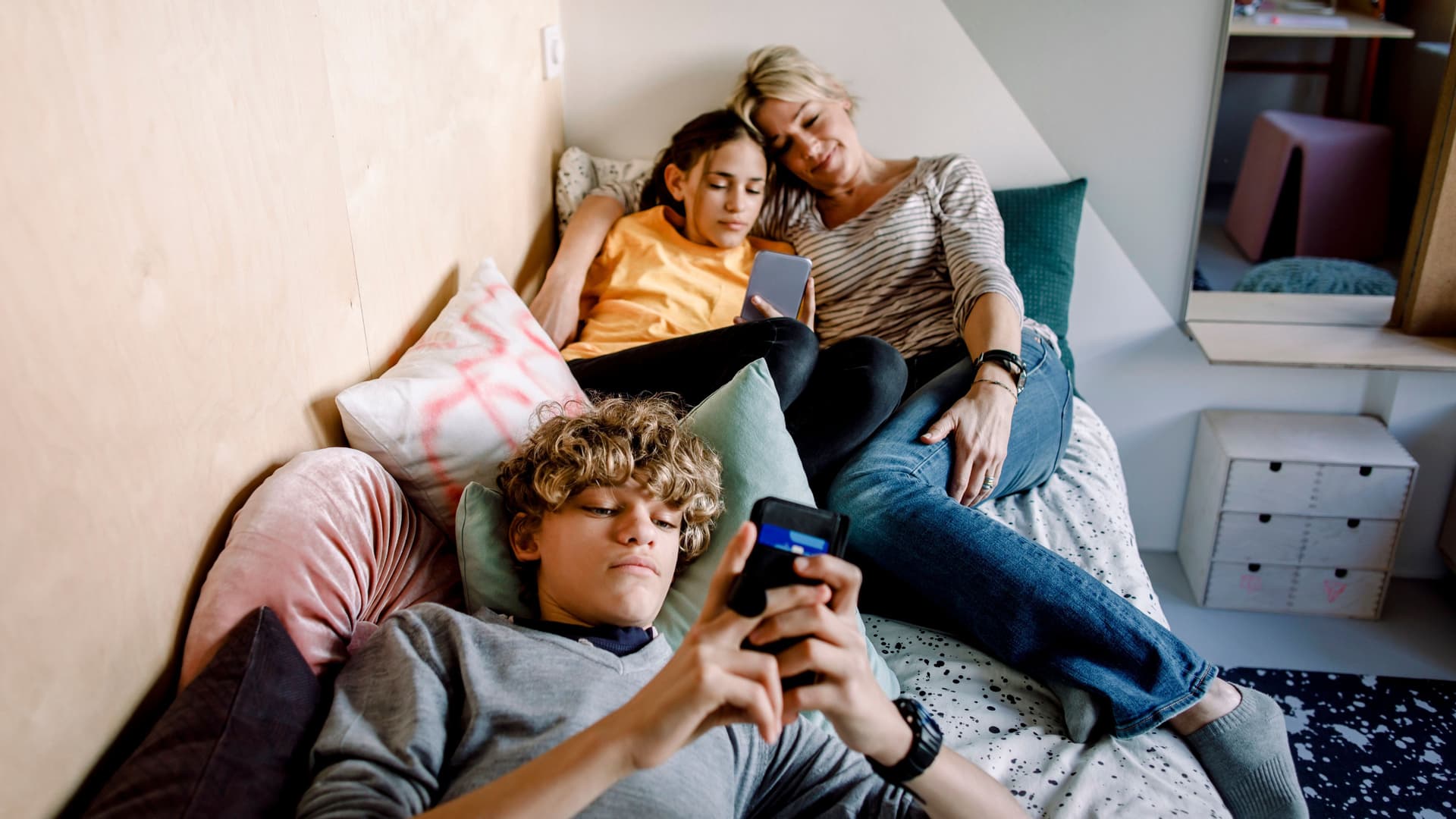 Mutter liegt mit Tochter und Sohn zusammen in einem Bett während diese am Handy sind.