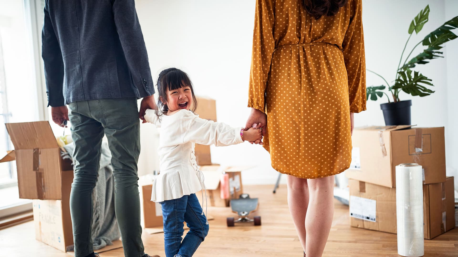 Lachende Kind an den Händen einer Frau und eines Mannes, die mit dem Rücken zur Kamera stehen. Sie stehen in einer Wohnung mit Umzugskisten.