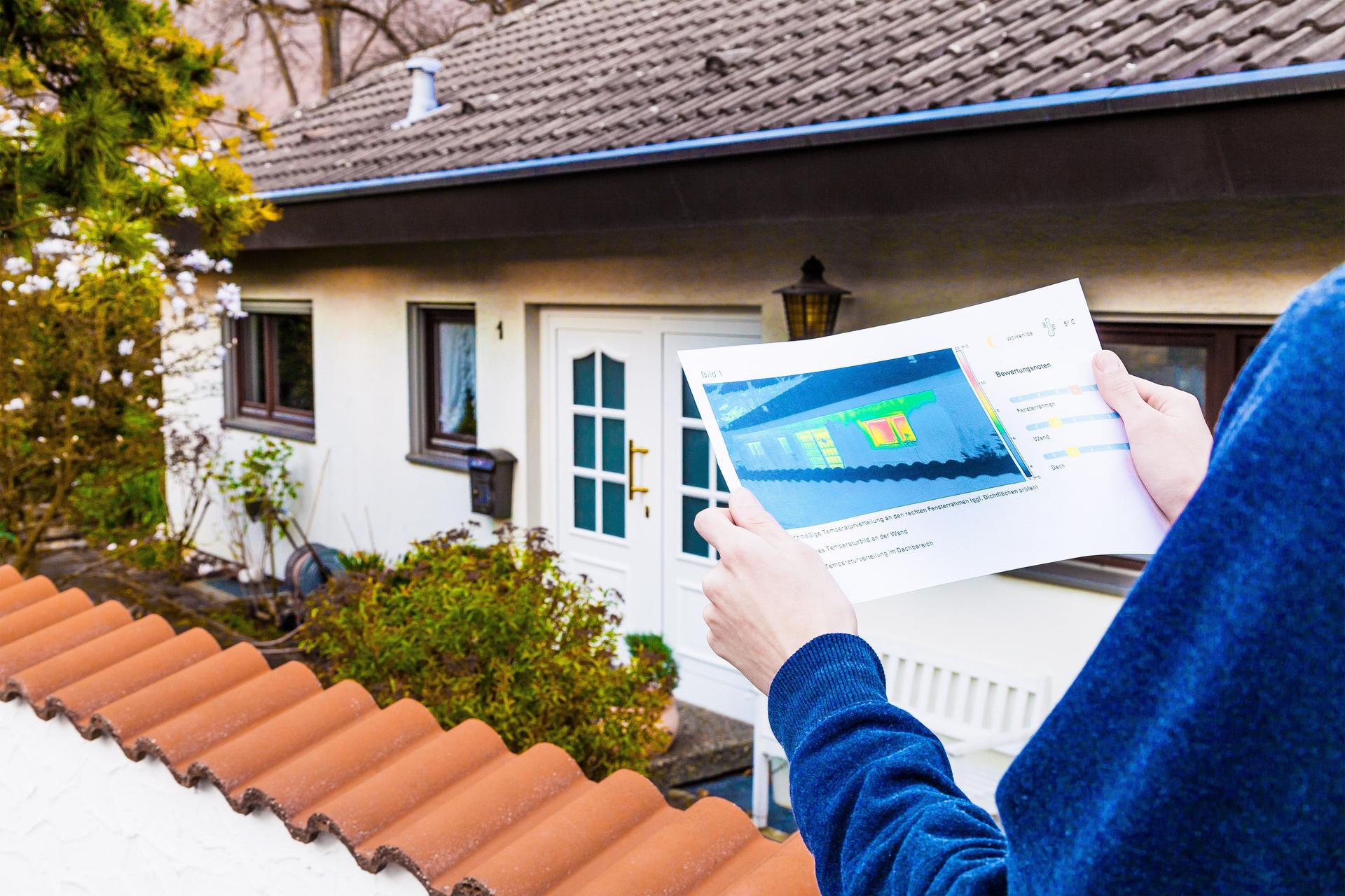 Eine Person hält die Thermografie eines Gebäudes in den Händen. Dahinter ist ein Haus zu sehen.