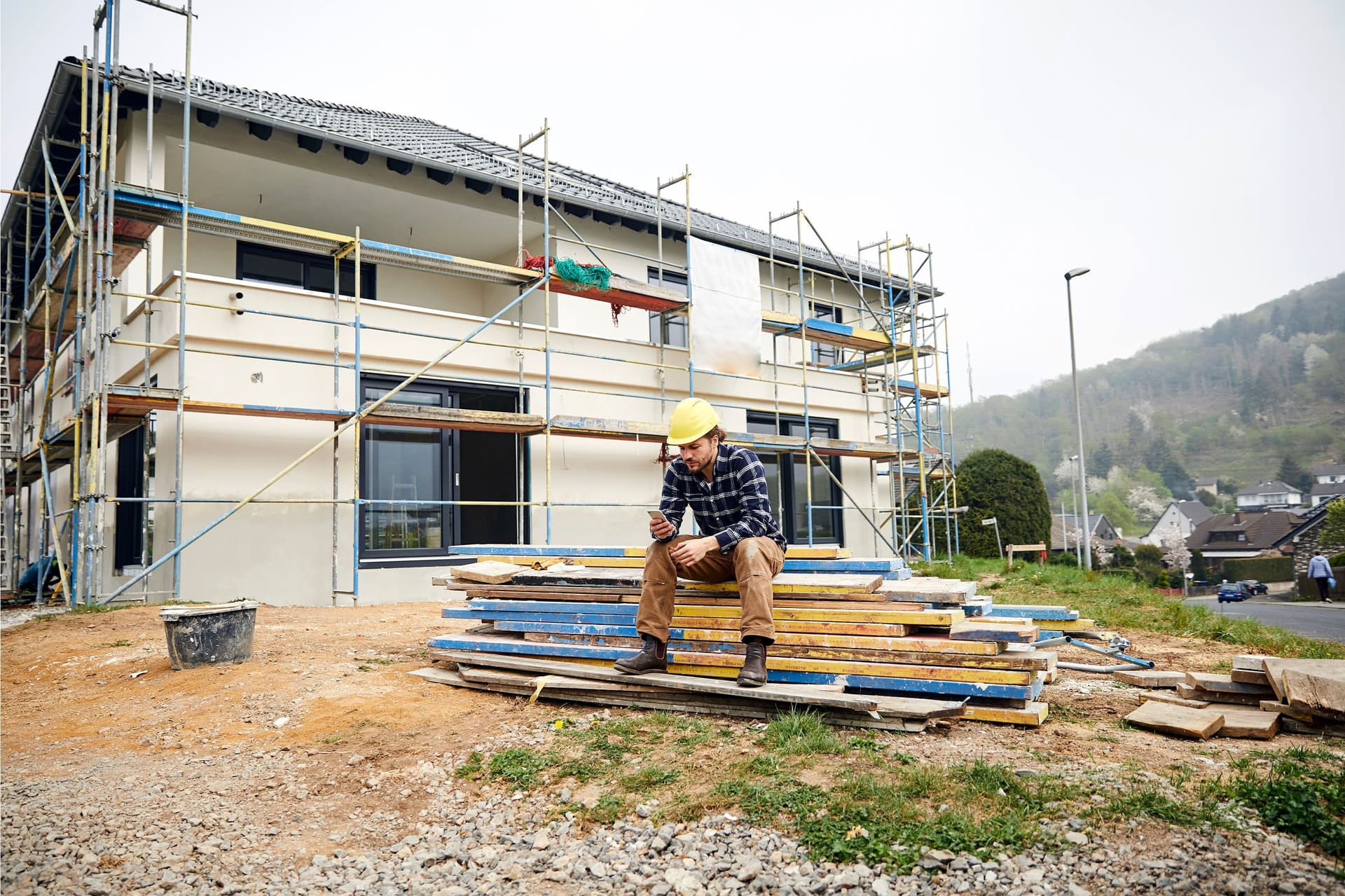 Ein Bauarbeiter mit Helm sitzt vor einer menschenleeren Baustelle auf einem Stapel Gerüstbrettern und schaut mit ernstem Gesichtsausdruck auf sein Smartphone.