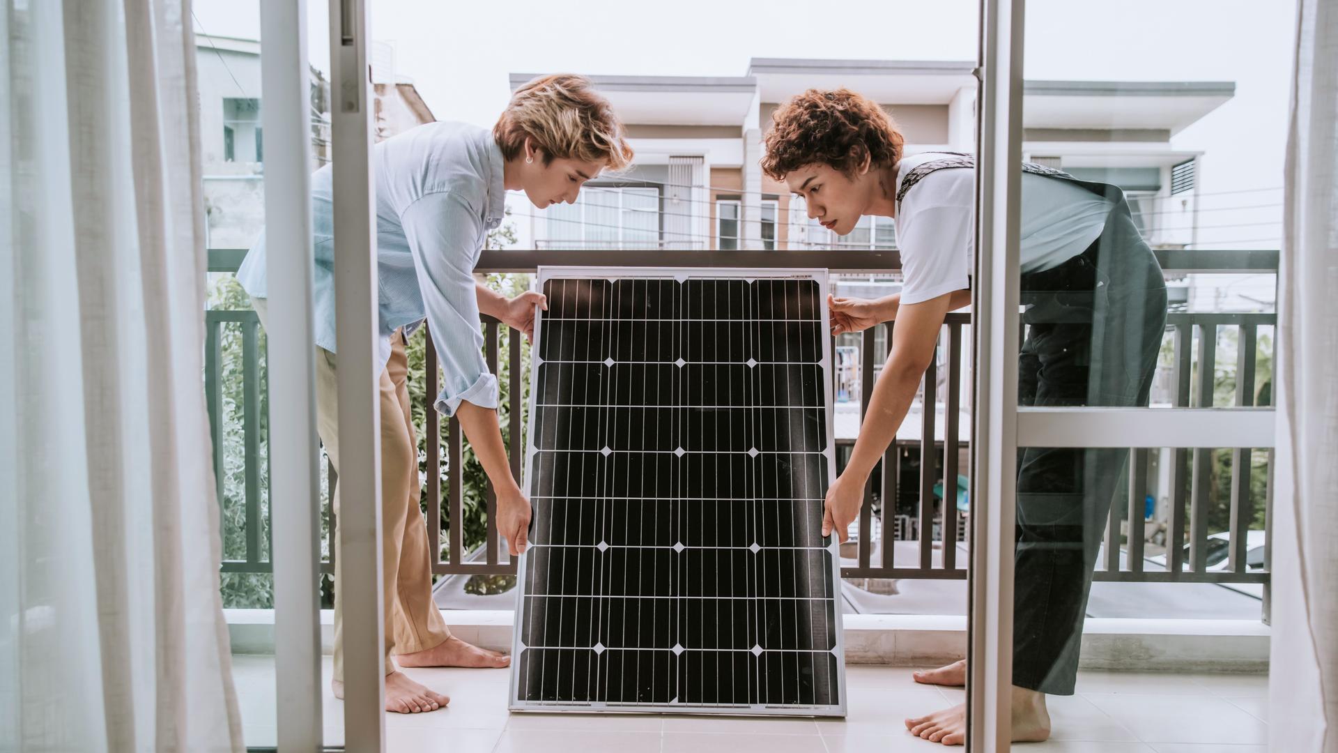 Zwei Personen stehen mit einem Solarpanel in den Händen auf dem Balkon eines Hauses.