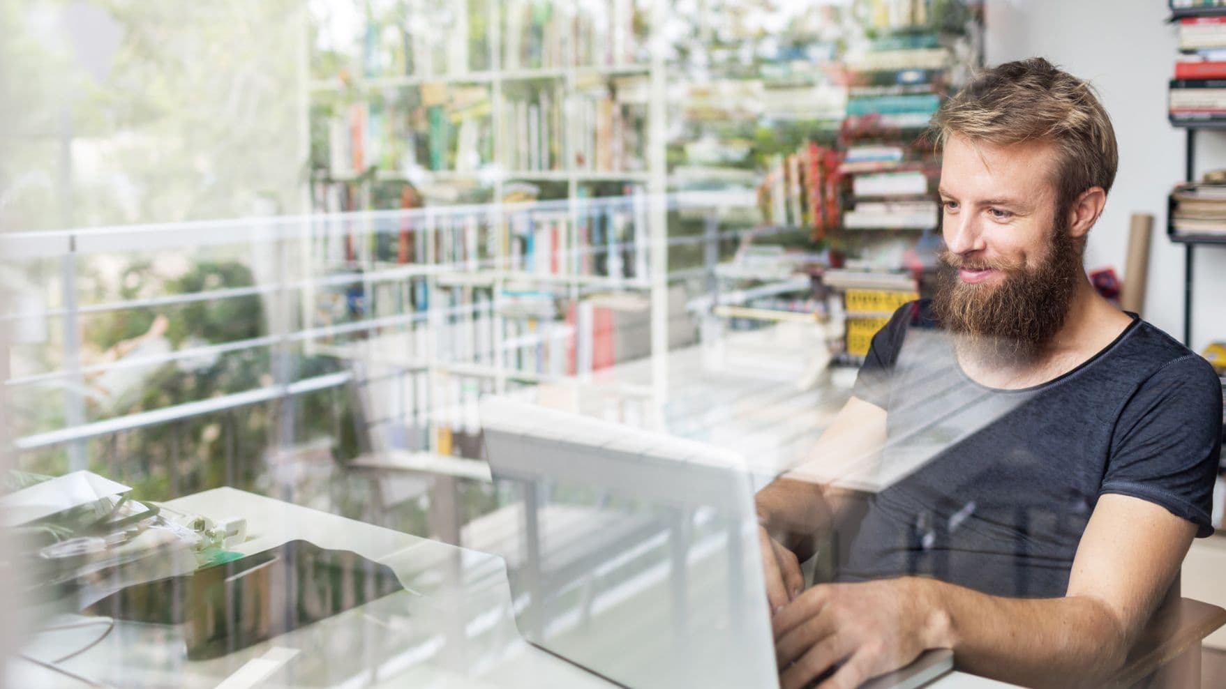 Ein bärtiger Mann arbeitet an einem Laptop vor einem Bücherregal, durch eine Fensterreflexion gesehen