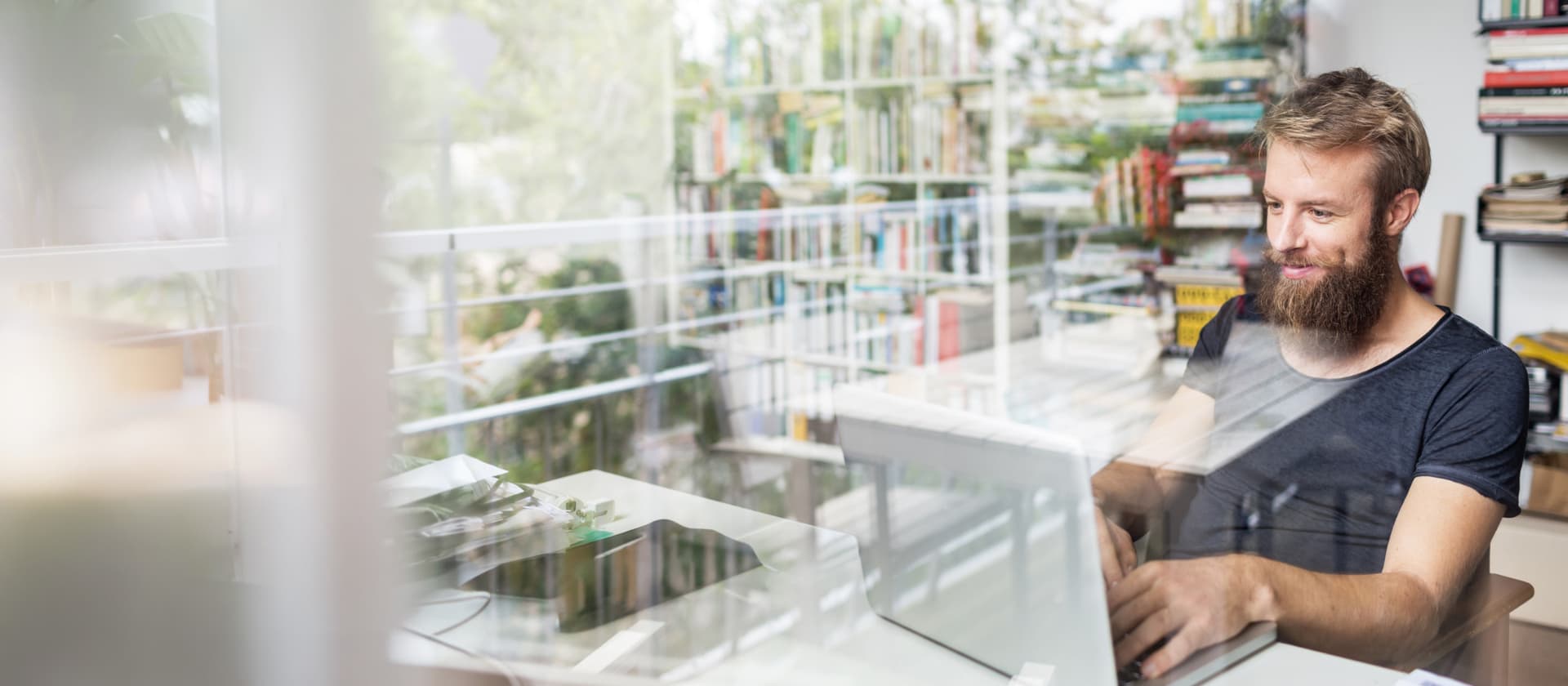 Ein bärtiger Mann arbeitet an einem Laptop vor einem Bücherregal, durch eine Fensterreflexion gesehen