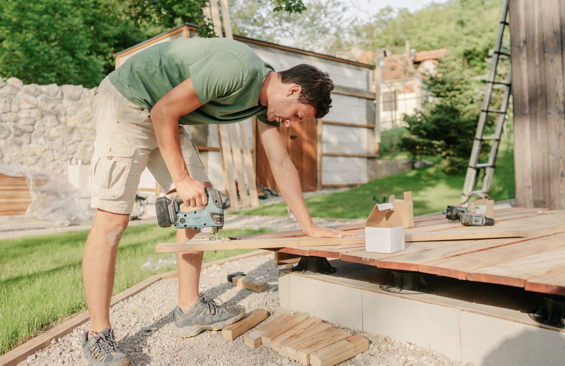 "Ein junger Mann, der sich im Freien in einem Garten befindet, bohrt mit einer elektrischen Bohrmaschine ein Loch in ein Brett. Er trägt Freizeitkleidung und ist konzentriert bei der Arbeit.