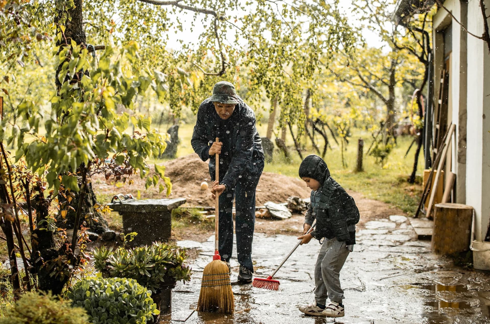"Ein Mann und ein Kind fegen gemeinsam bei leichtem Regen eine Terrasse. Beide tragen Regenjacken, während sie mit Besen die nasse Oberfläche säubern. Im Hintergrund sind Gartenmöbel und Pflanzen zu sehen.
