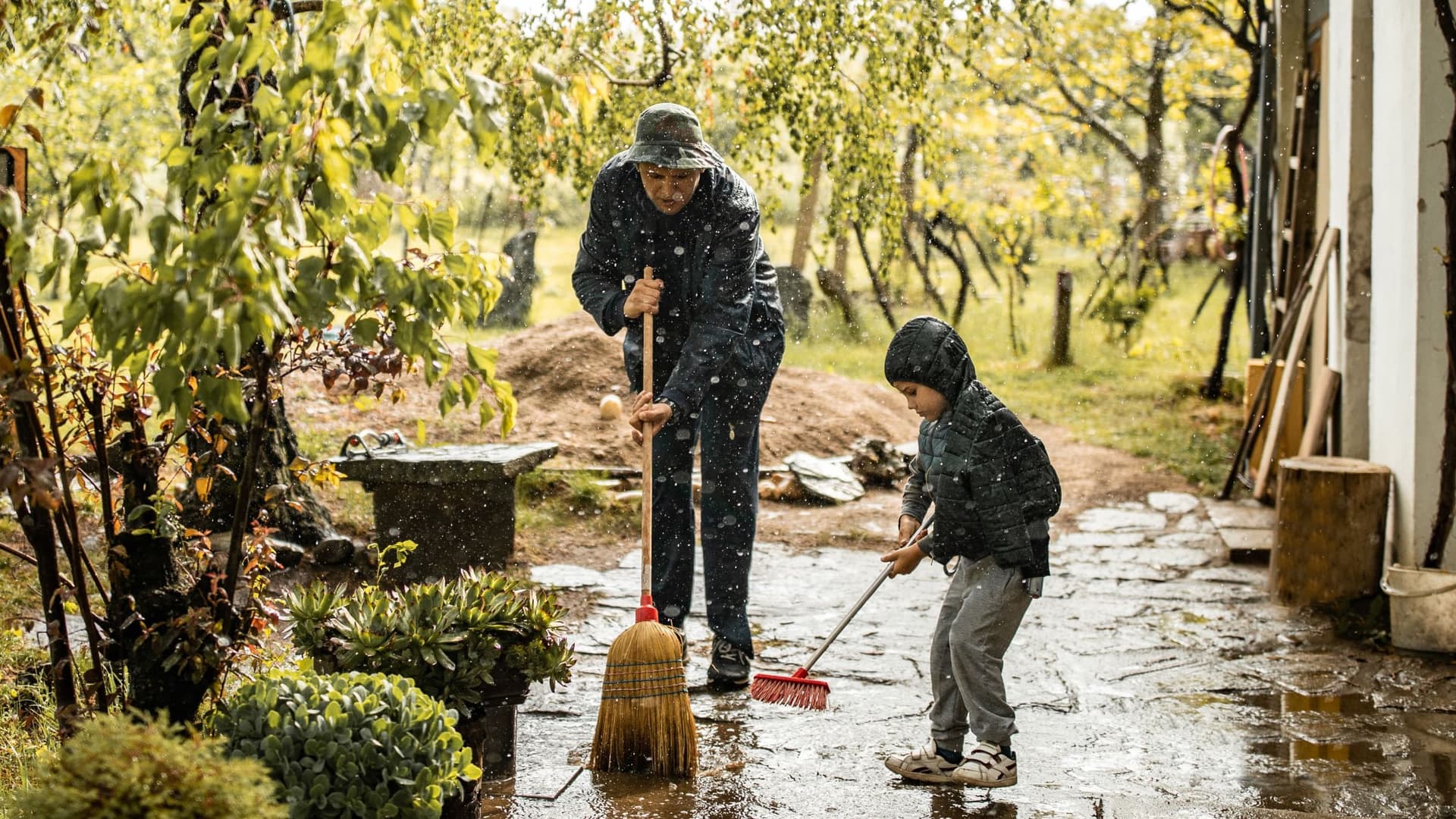 Ein Mann und ein Kind fegen gemeinsam bei leichtem Regen eine Terrasse. Beide tragen Regenjacken, während sie mit Besen die nasse Oberfläche säubern. Im Hintergrund sind Gartenmöbel und Pflanzen zu sehen.