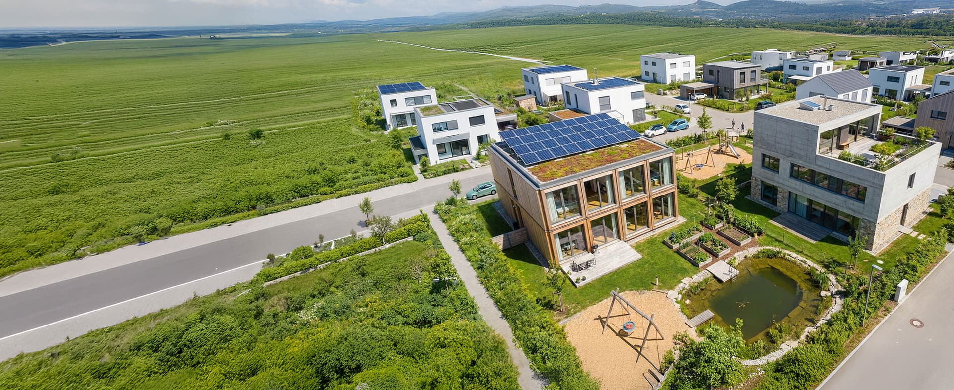 Vogelperspektive auf moderne Wohnhäuser mit Solarzellen, Dachbegrünung, Gemüsegarten, Spielplatz und natürlichem Teich in einer Wohnsiedlung.