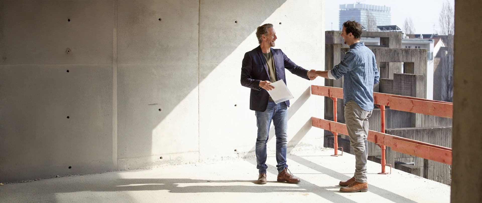 Zwei Männer auf einer Baustelle geben sich die Hand, einer hält Bauunterlagen in der Hand