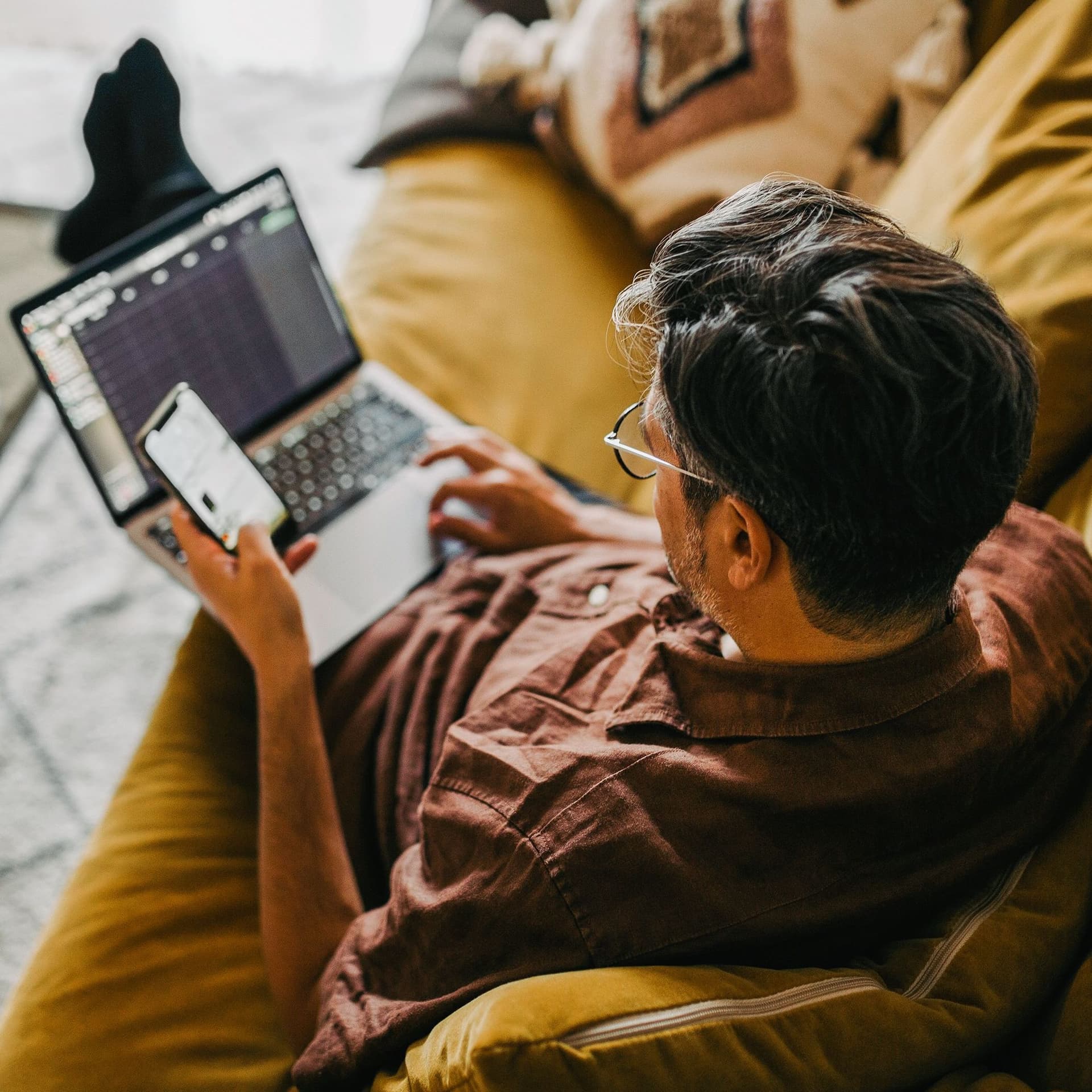 Blick von oben auf einen Mann, der entspannt mit Laptop auf dem Schoß auf dem Sofa sitzt. Er schaut auf sein Smartphone.