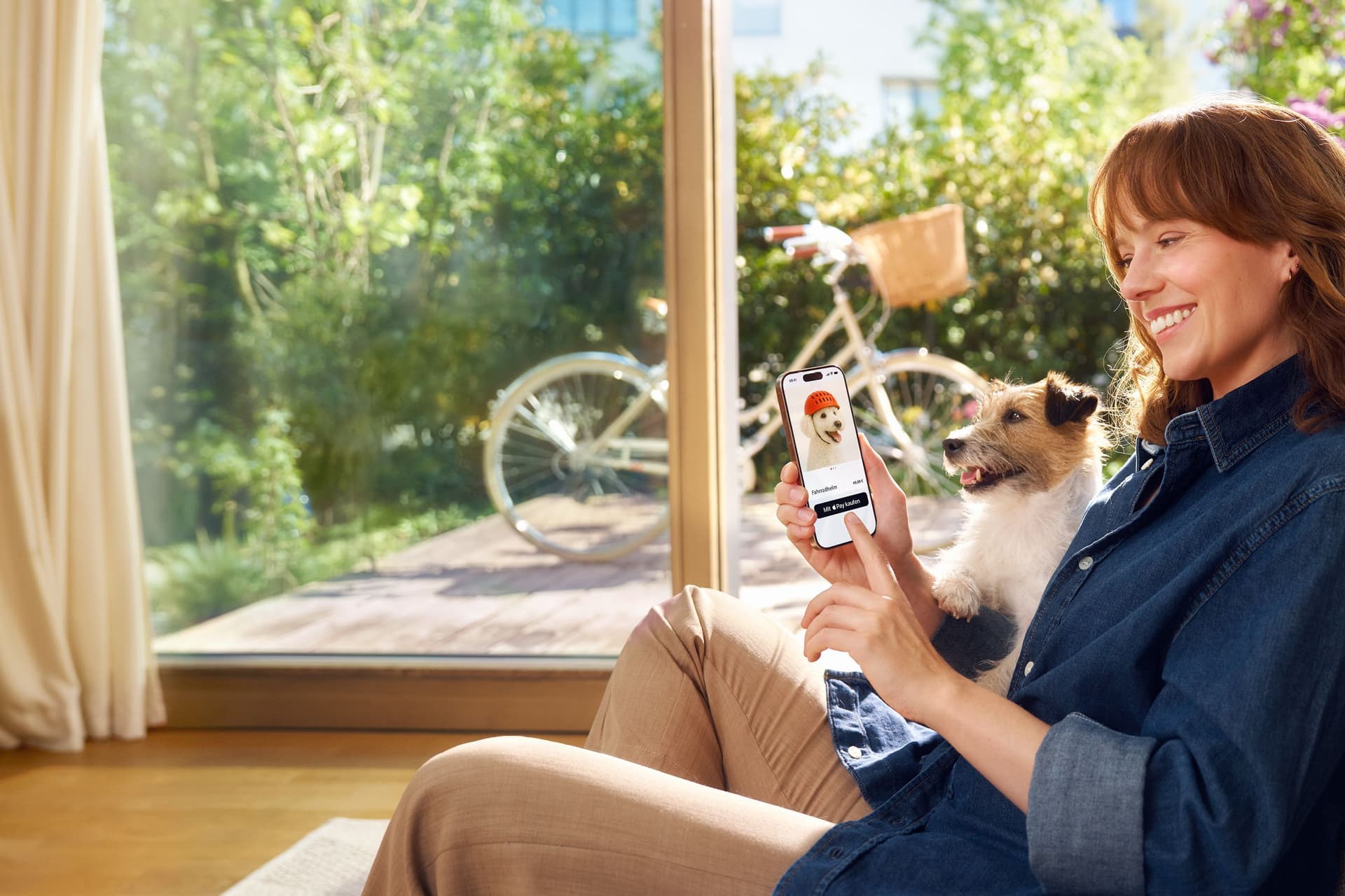 Eine Frau sitzt lächelnd mit einem Hund auf dem Schoß auf dem Sofa und nutzt ihr Smartphone, um online einen Fahrradhelm für Hunde zu kaufen. Im Hintergrund ist ein sonniger Garten mit einem Fahrrad zu sehen.