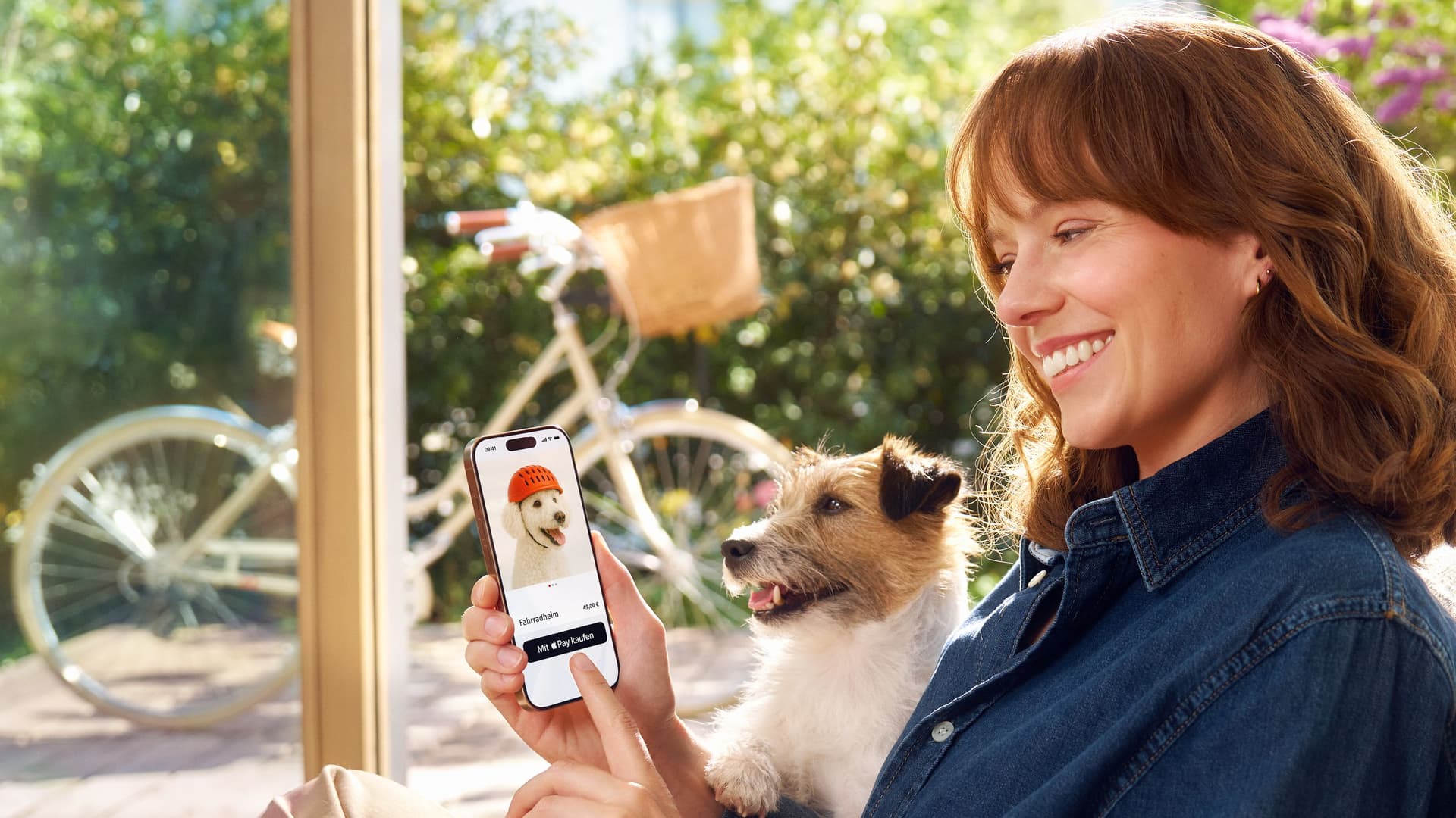 Eine Frau sitzt lächelnd mit einem Hund auf dem Schoß auf dem Sofa und nutzt ihr Smartphone, um online einen Fahrradhelm für Hunde zu kaufen.