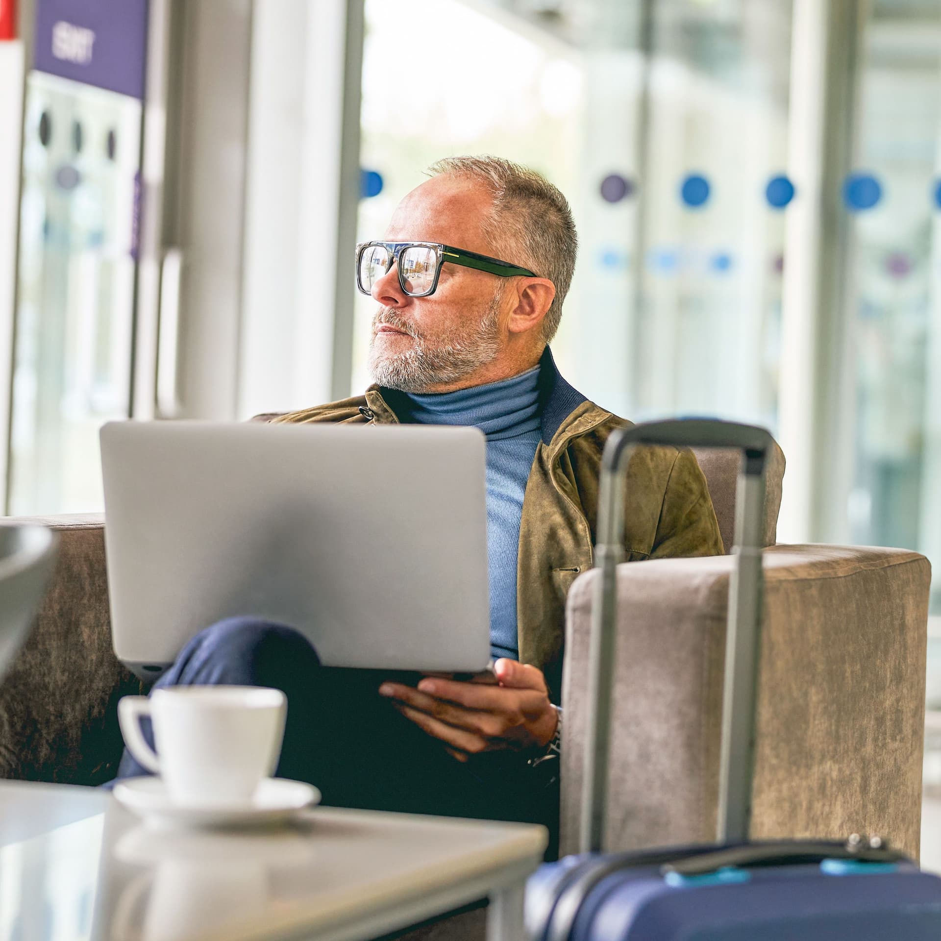 Älterer Herr sitzt mit Laptop auf dem Schoß vor Glasfront in einem Lounge-Bereich und schaut raus.