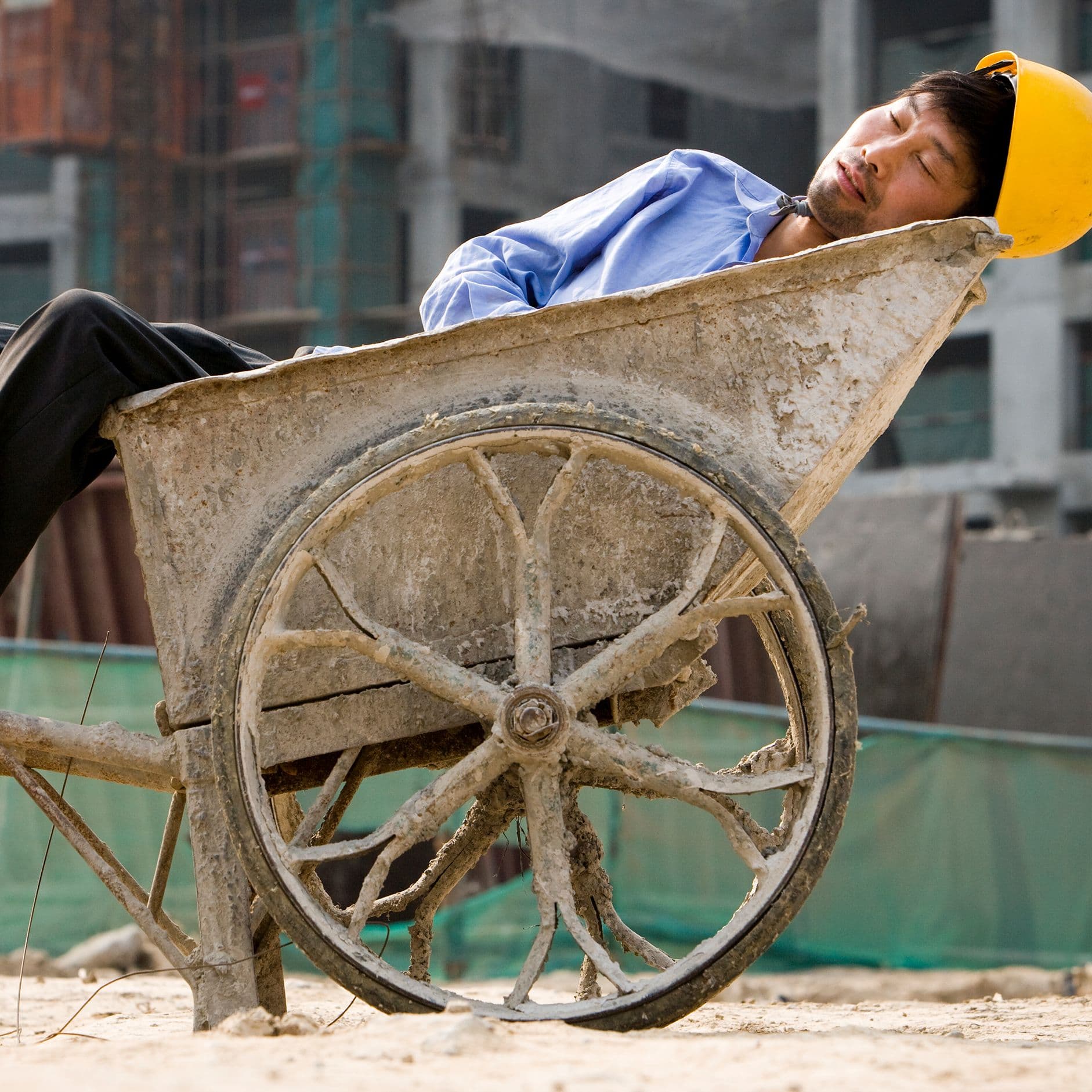 Ein Bauarbeiter mit gelbem Schutzhelm schläft erschöpft in einer stark verschmutzten Schubkarre auf einer Baustelle. Im Hintergrund sind unscharfe Gebäudestrukturen zu erkennen, die die raue Arbeitsumgebung unterstreichen.
