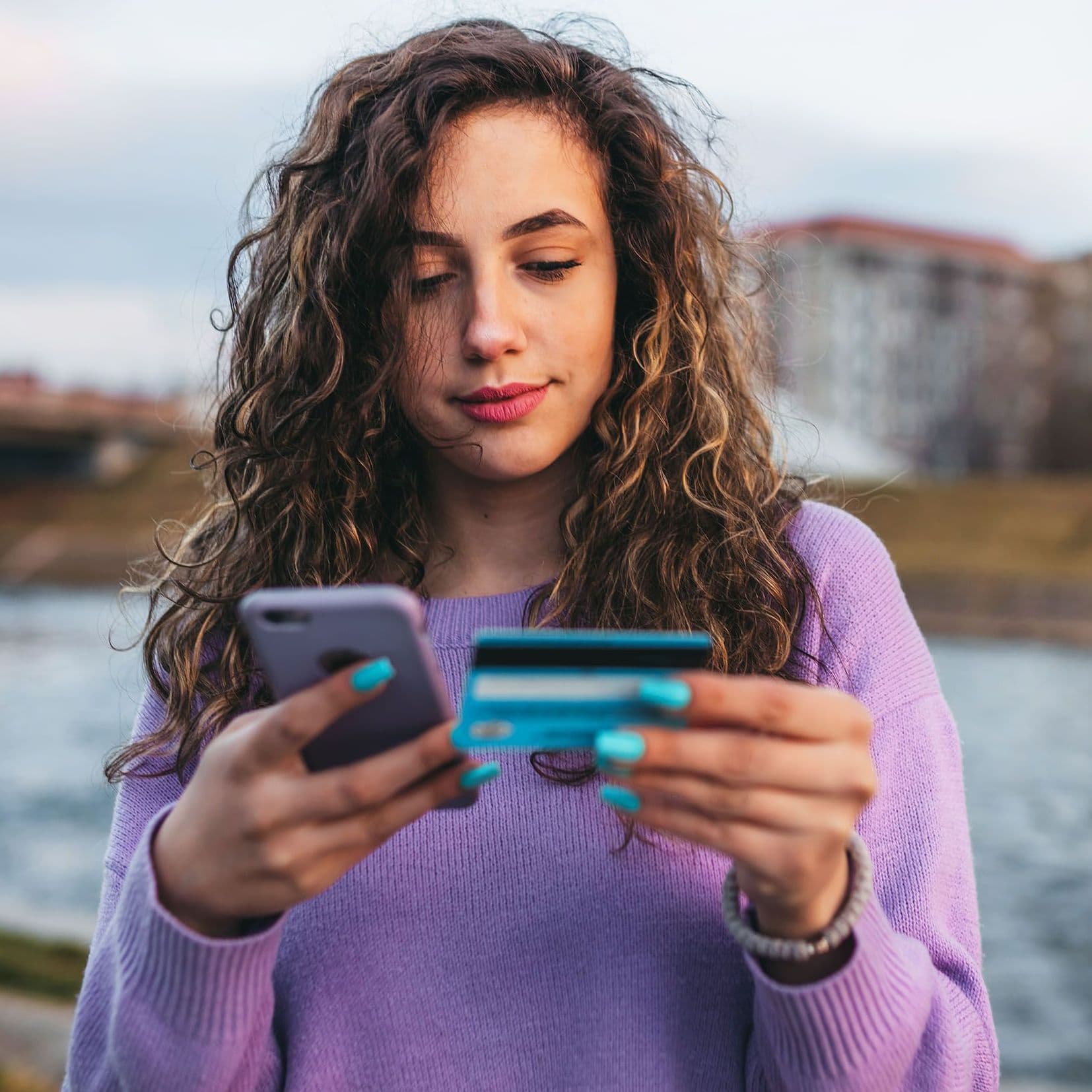 Eine junge Frau mit langen lockingen, braunen Haaren in einem violetten Pullover schaut auf ihr Smartphone und hält ihre Geldkarte in der Hand.
