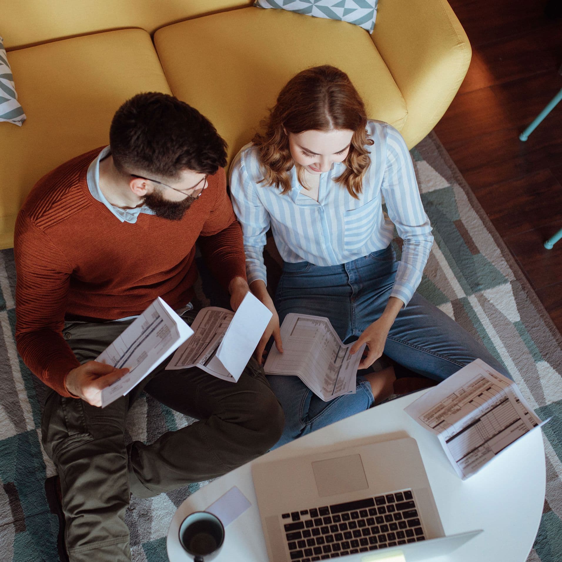 Paar sitzt auf dem Boden vor Sofa und prüft gemeinsam Rechnungen und Unterlagen mit Laptop auf dem Tisch