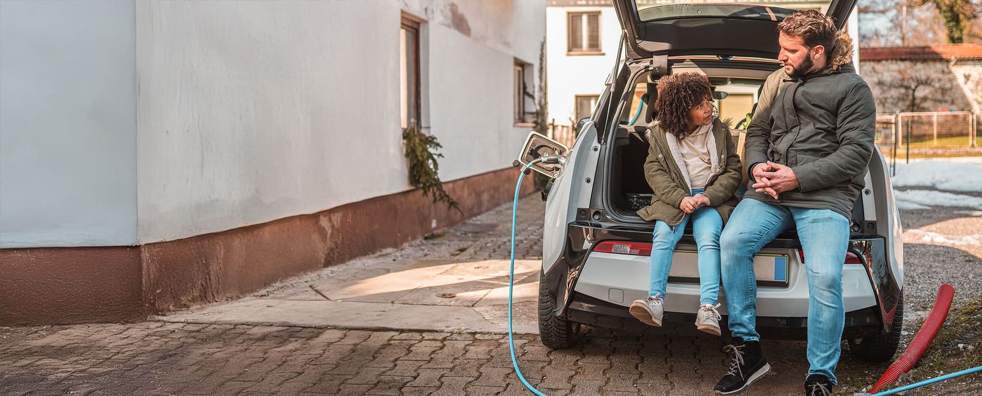 Vater und Kind sitzen im offenen Kofferraum eines Elektroautos, das vor einem Haus lädt
