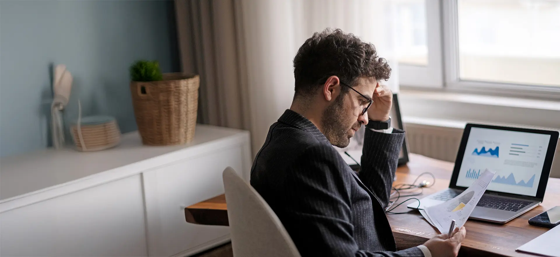 Mann mit Brille sitzt am Schreibtisch und schaut konzentriert auf Finanzdokumente und Laptop mit Diagrammen.