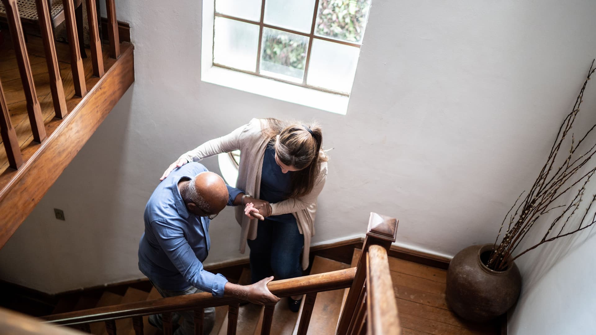 Eine junge Frau hilft einem älteren Mann in einem alten Treppenhaus aus Holz die Treppe hoch