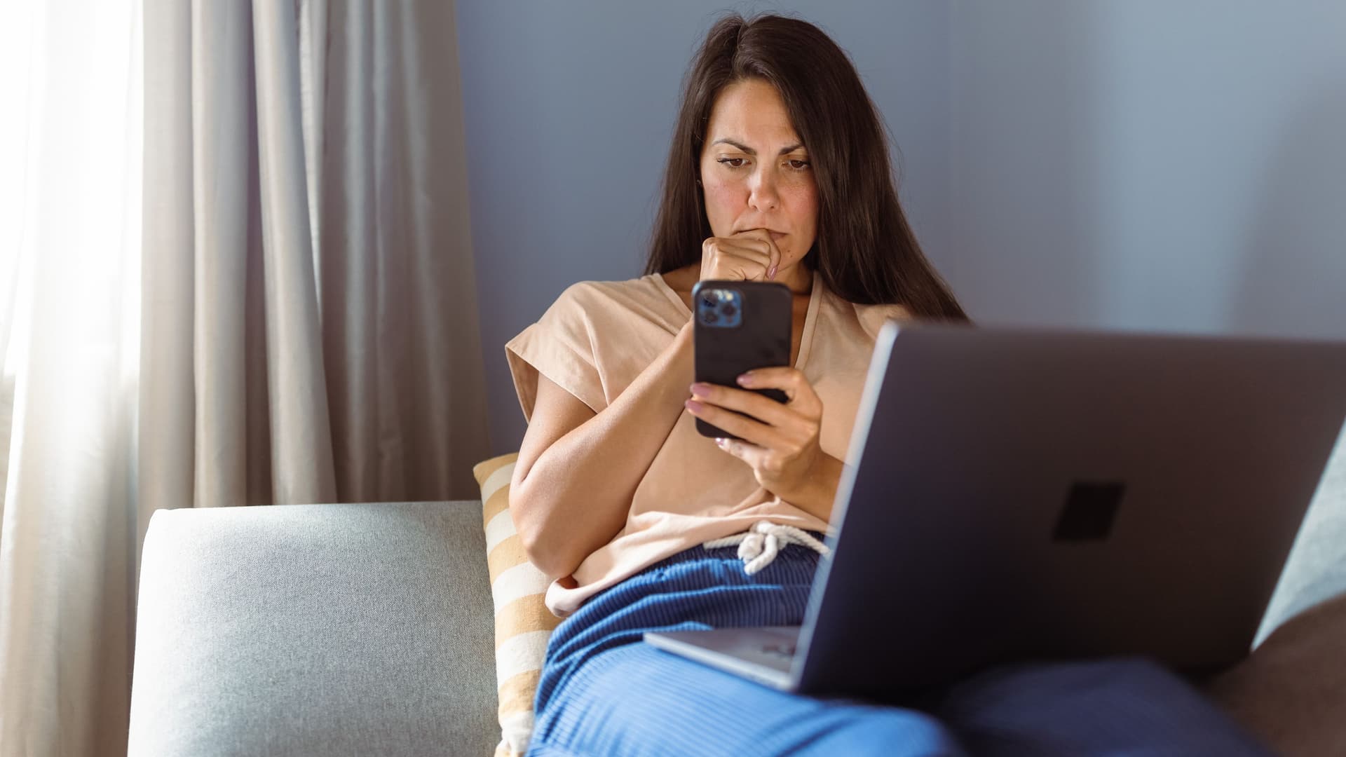 Frau auf Sofa mit Laptop, hält ein Smartphone und wirkt nachdenklich