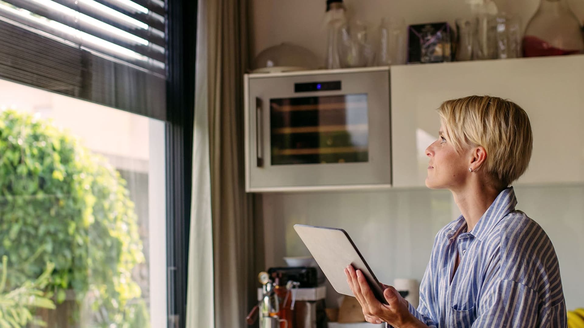 Eine Frau bedient elektrische Rollläden in einem modernen Wohnraum. Sie steht vor einem Fenster und zieht an einem Schalter, während die Rollläden sanft nach unten fahren.